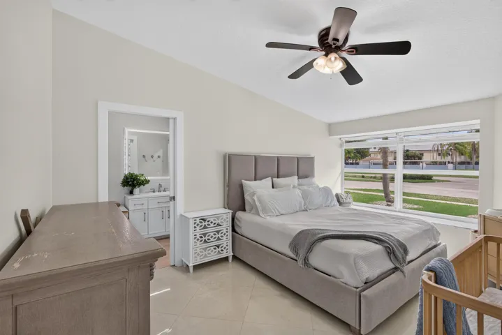 Spacious primary bedroom featuring a gray upholstered bed frame, wood-finish nightstand with decorative hardware, ceiling fan with integrated lighting, and expansive glass doors opening to outdoor space