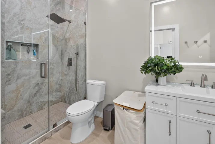 Spa-inspired primary bathroom featuring a frameless glass-enclosed walk-in shower with marble-look tile surround and integrated niche, dual-sink vanity with white cabinetry and brushed nickel hardware, LED-backlit mirror, and porcelain tile flooring throughout