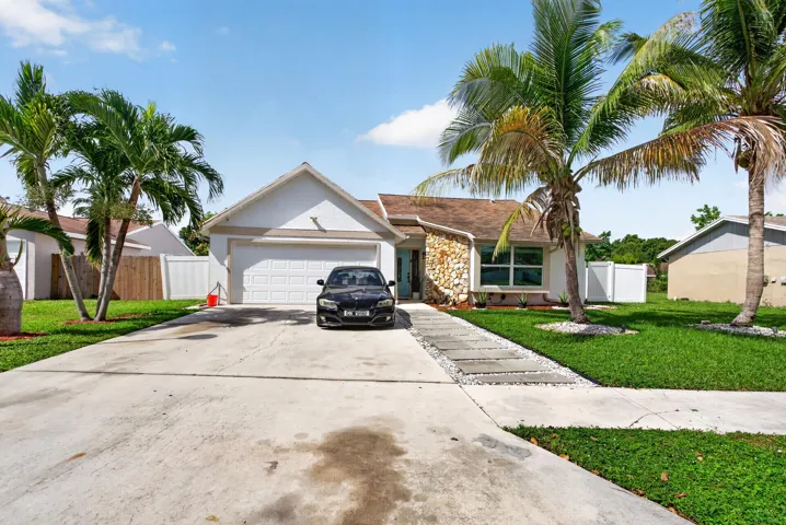 Single-story residence featuring a two-car garage with driveway access, gabled roofline with stone and stucco exterior detailing, mature palm landscaping, and manicured lawn