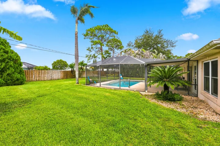 Expansive backyard featuring a screened-in swimming pool, a covered patio area, a manicured lawn, mature palm trees, and a wooden perimeter fence