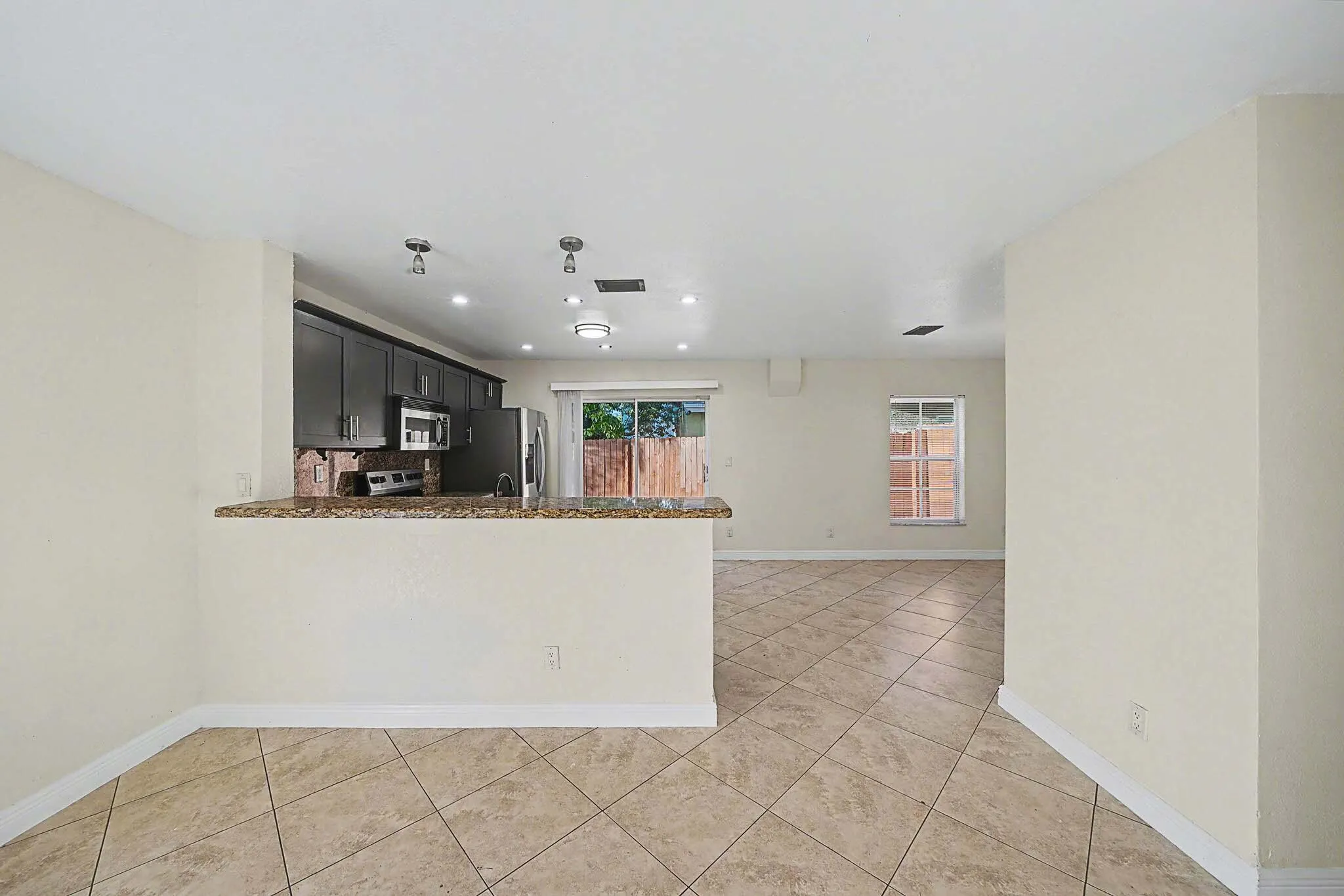 Open-concept living area with neutral tile flooring, a kitchen featuring dark cabinetry, stainless steel appliances, and a granite-look countertop peninsula