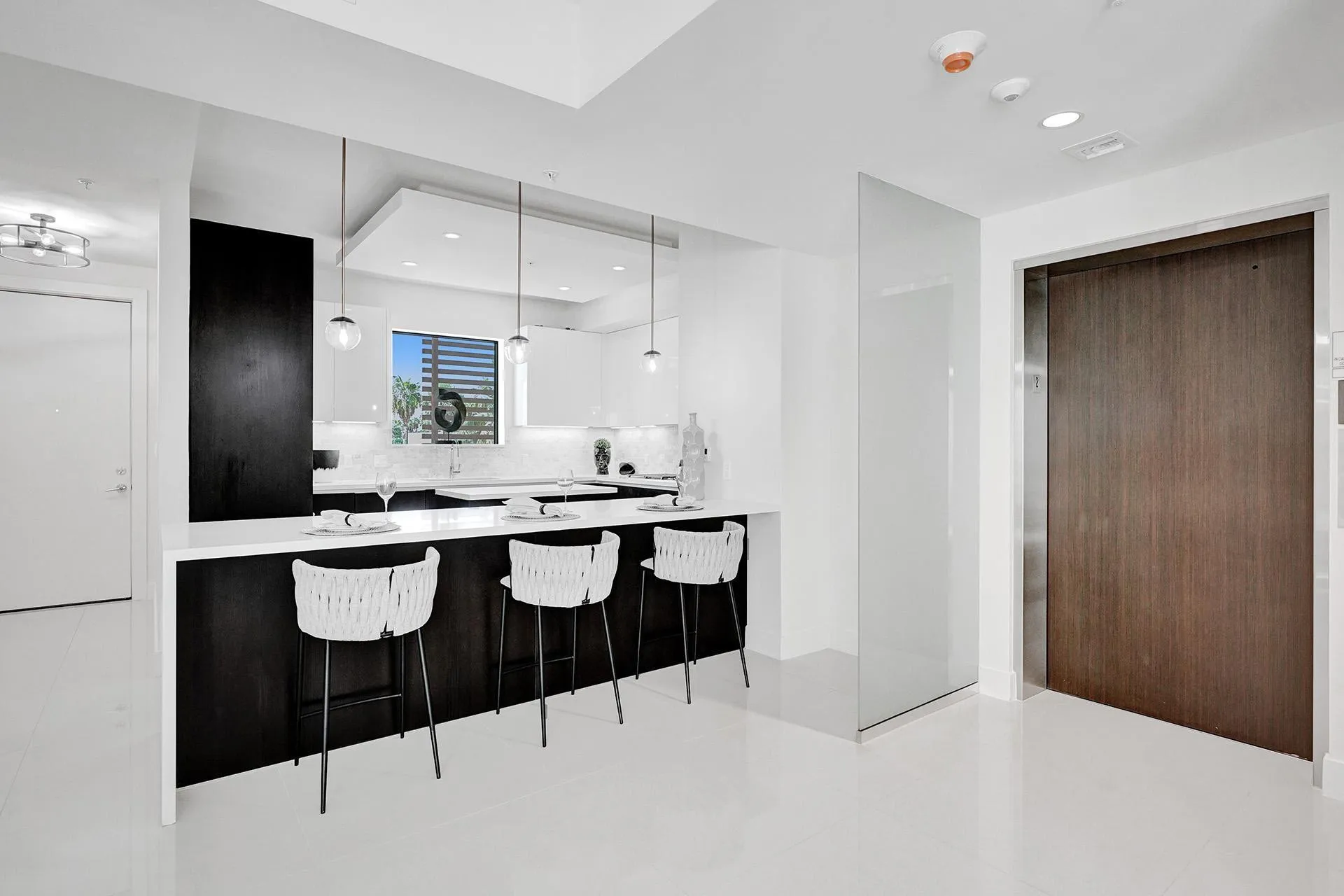 Modern kitchen featuring an extended white countertop, dark wood-finish cabinetry, globe pendant lighting, and a window with horizontal blinds