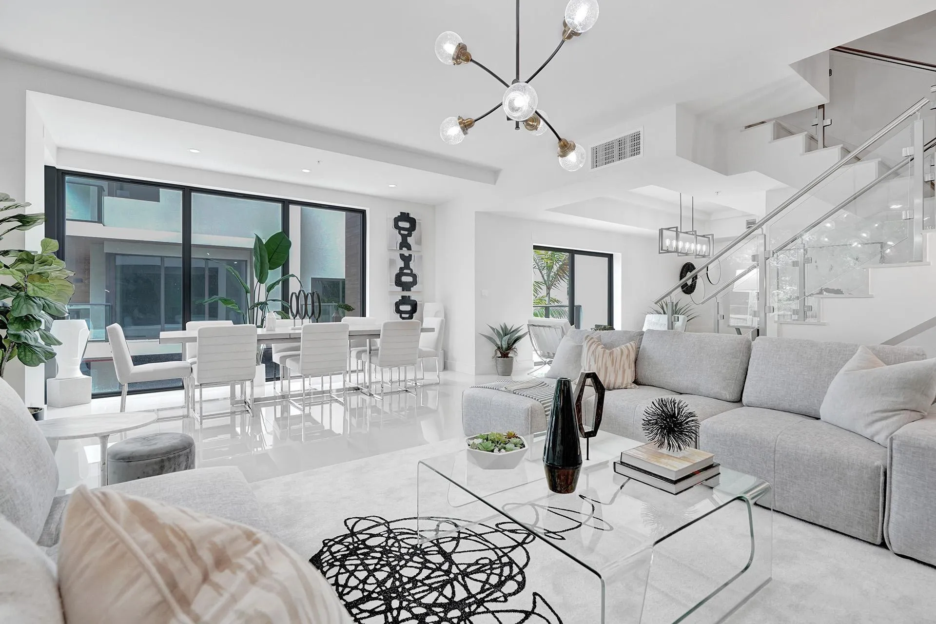 Spacious open-concept living area featuring a modern chandelier, polished tile flooring, and a glass-paneled staircase with a metal handrail