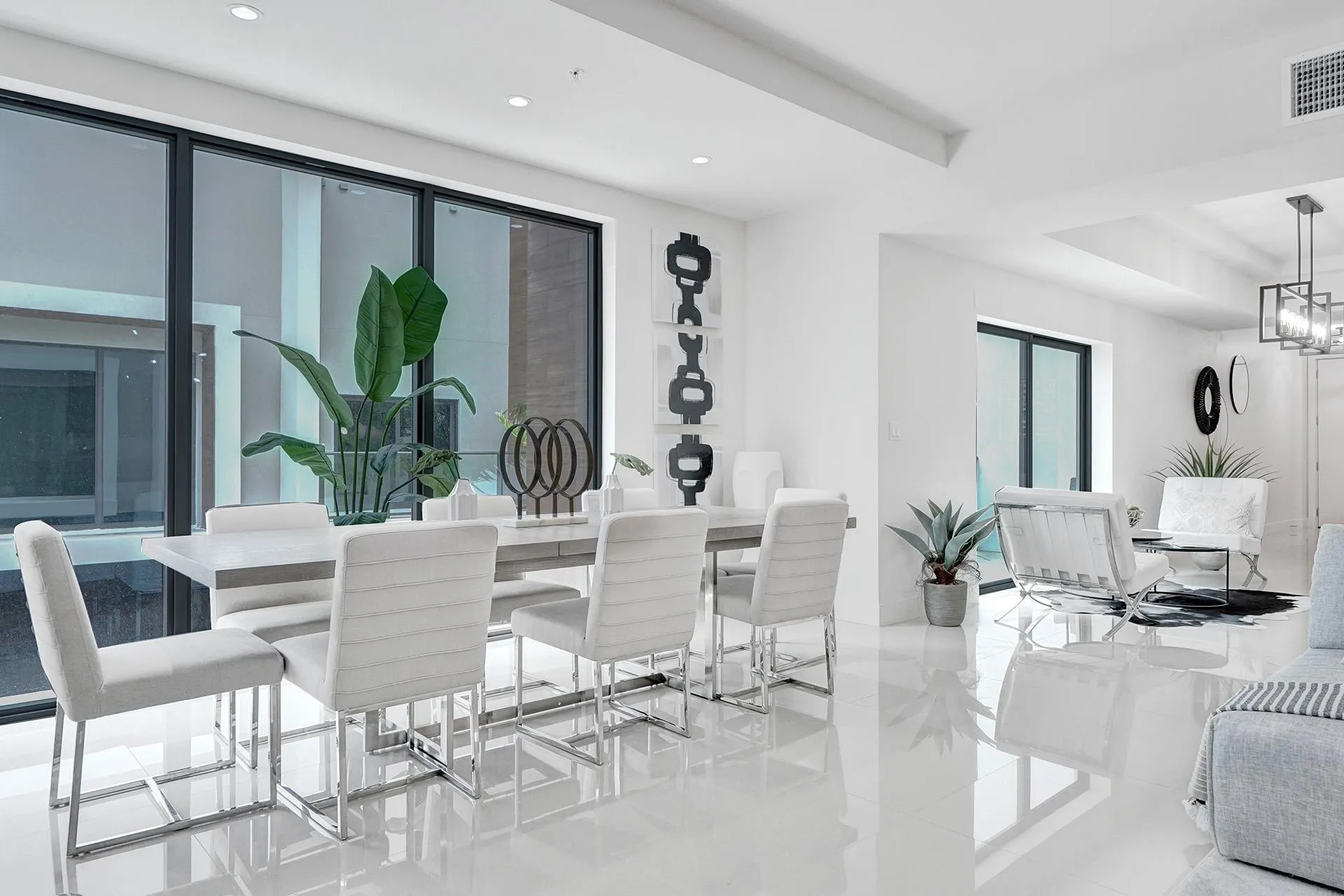 Sophisticated open-concept dining and living area featuring polished light-toned flooring, floor-to-ceiling windows with dark-framed glazing, contemporary white upholstered dining chairs with metal frames, and a sleek white dining table