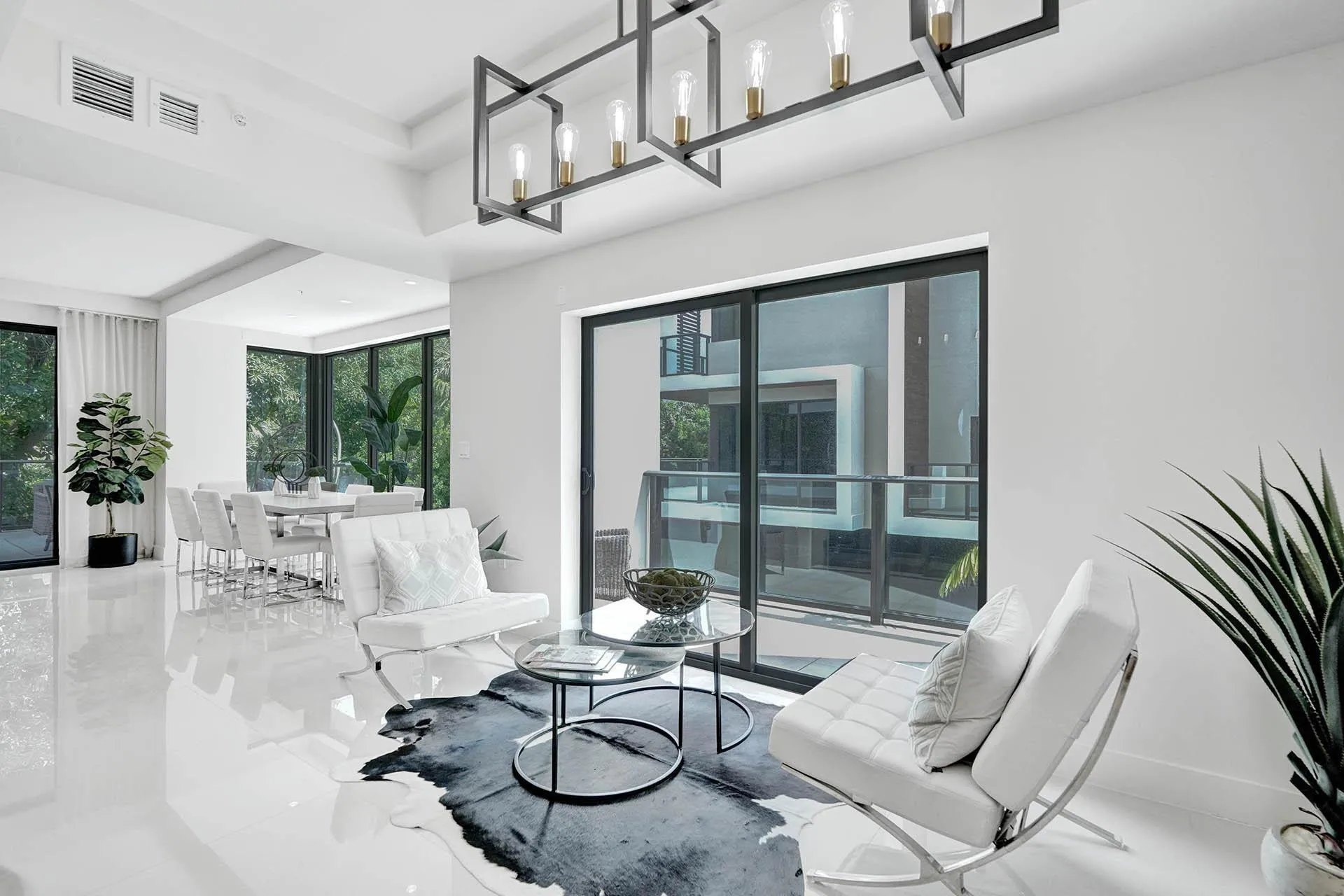 Spacious living area featuring polished white flooring, a modern linear chandelier with exposed bulbs, and a sliding glass door opening to a private balcony