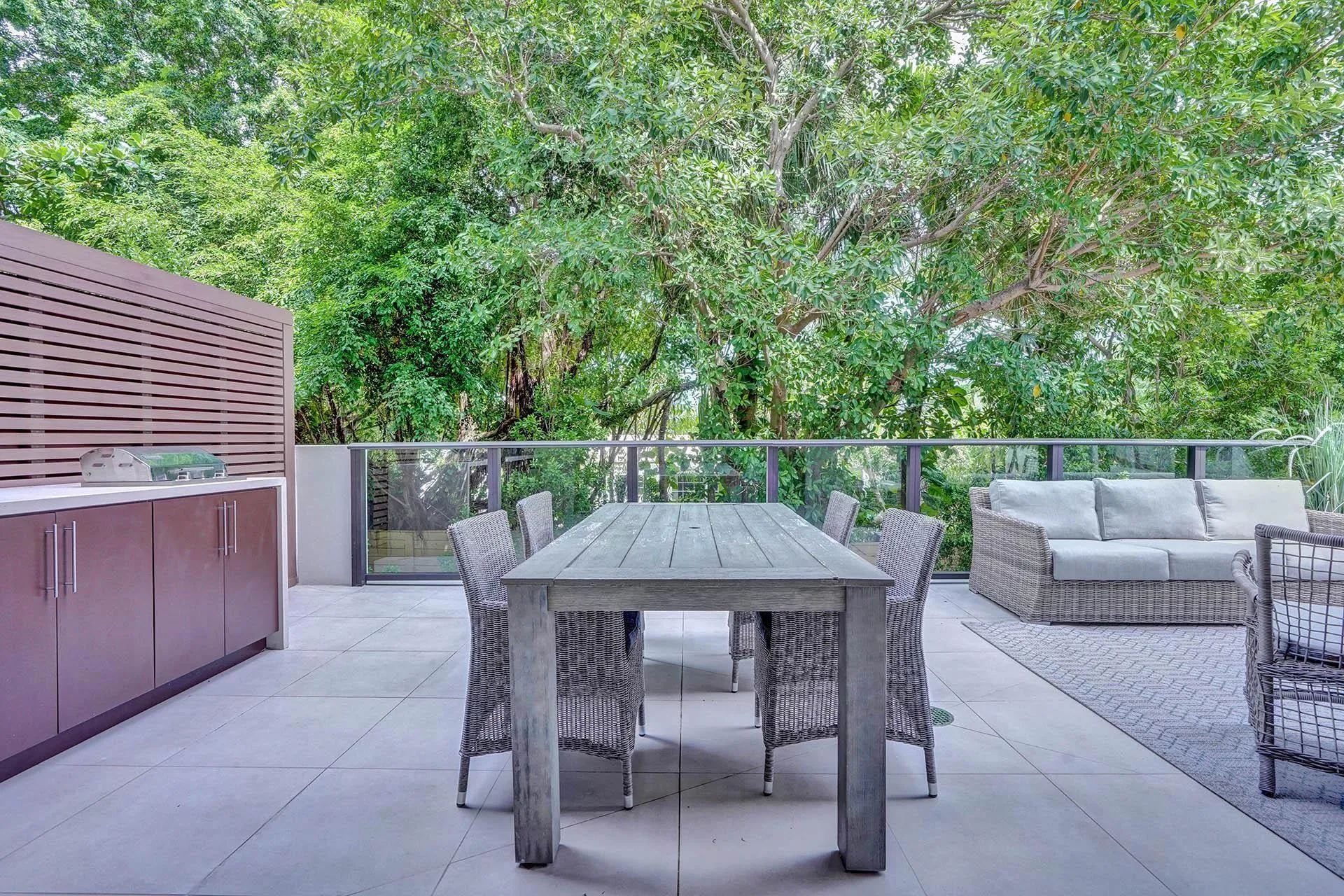Expansive outdoor terrace featuring light-toned large format tile flooring, a horizontal slat privacy screen, and a built-in outdoor kitchen with brown cabinetry and a light countertop