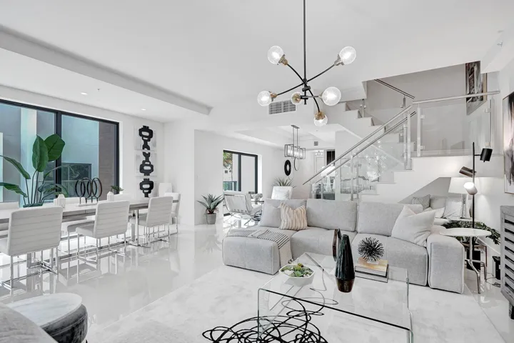 Modern open-concept living space featuring polished light-toned flooring throughout, floor-to-ceiling windows with natural light, a sculptural multi-pendant ceiling fixture, a floating staircase with glass railings, and contemporary furnishings arranged across the expansive great room