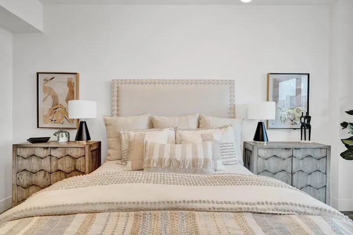 Serene primary bedroom featuring an upholstered headboard with nailhead trim, layered bedding in neutral tones, matching nightstands with geometric patterning, table lamps with black bases, and artwork accents on either side