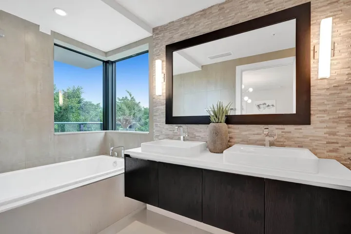 Bathroom featuring a built-in soaking tub, corner window, dual vessel sinks, floating vanity with dark wood-finish cabinetry, and a full-width mirror with sconce lighting
