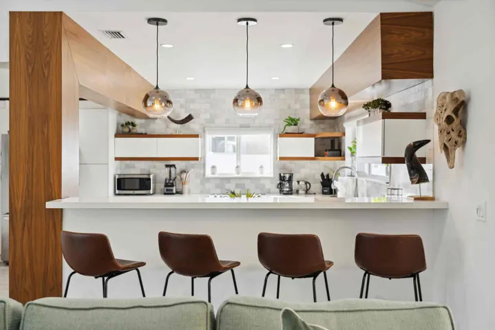 Kitchen featuring a white countertop island with integrated seating, three pendant lights, wood-finish beams, white cabinetry with wood-finish accents, and a tiled backsplash
