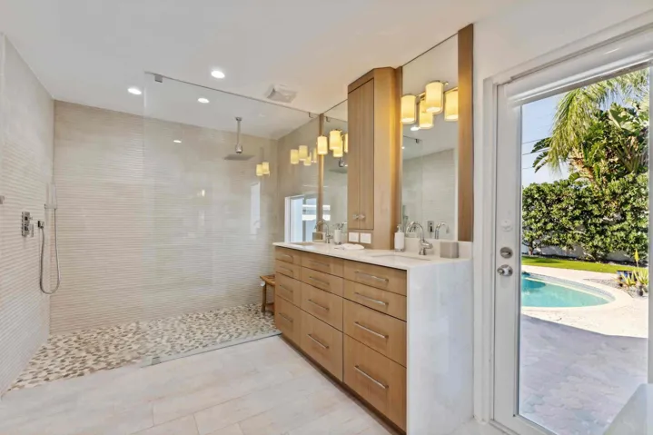 Spa-inspired primary bathroom featuring a spacious walk-in shower with white subway tile surround and pebble stone flooring, custom wood-finish vanity with dual sinks, integrated mirror lighting with warm-toned pendant fixtures, and direct access to the backyard through glass sliding doors