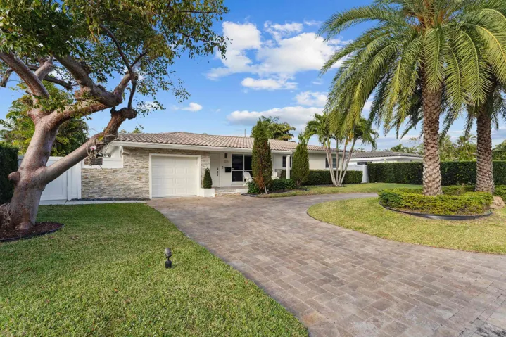 Elegant single-story residence featuring a stone and stucco exterior, wide circular driveway with paver construction, mature landscaping including a striking specimen tree and lush palm plantings, manicured lawn, and professional hedge borders framing the property