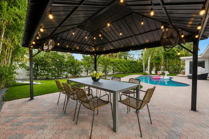 Elegant covered patio featuring a black metal gable roof with integrated pendant lighting and ceiling fans, anchoring a spacious brick-paved entertaining area