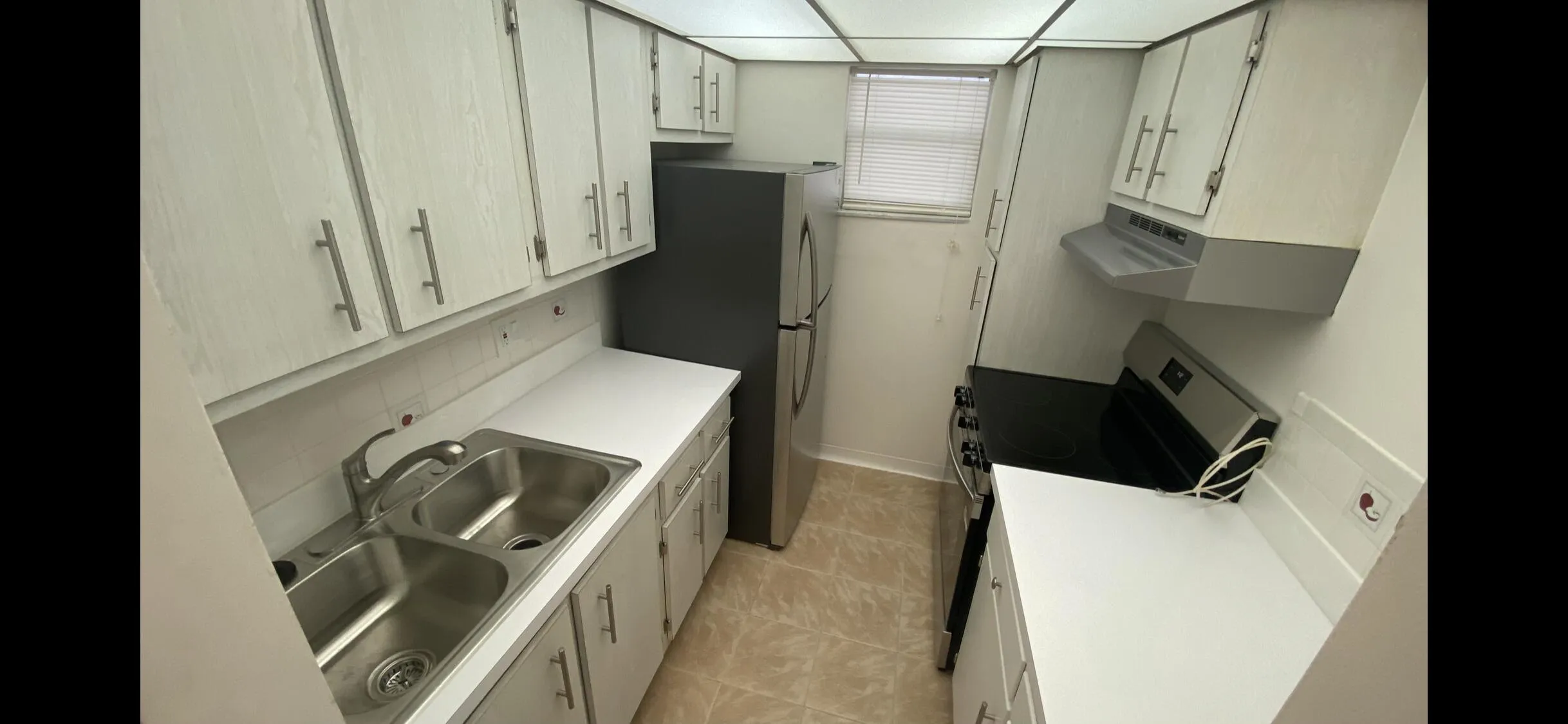 Galley kitchen featuring white cabinetry, light countertops, dual basin stainless steel sink, tiled flooring, and a window