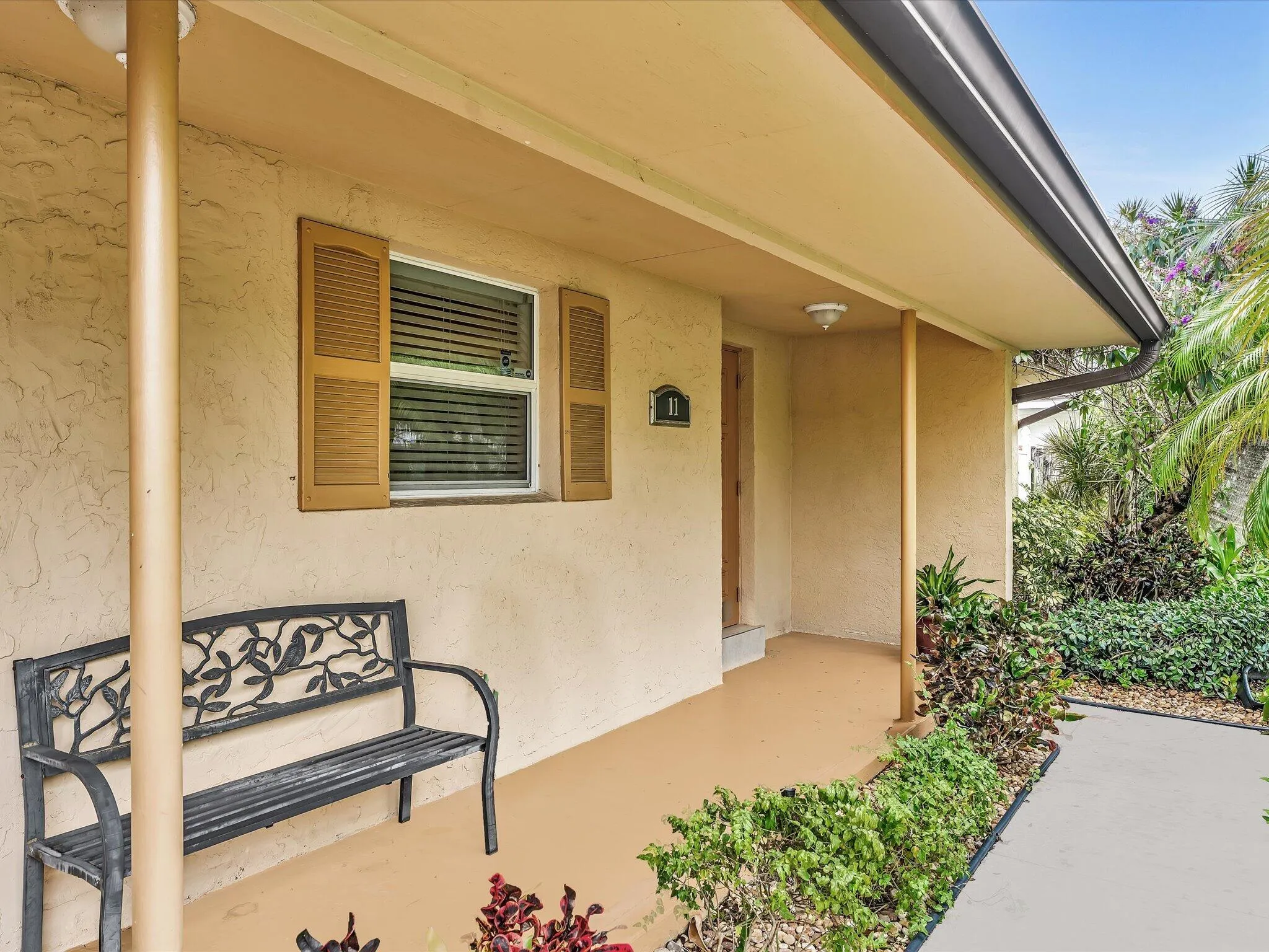 Covered entryway with textured stucco exterior, window with louvered shutters, and mature landscaping