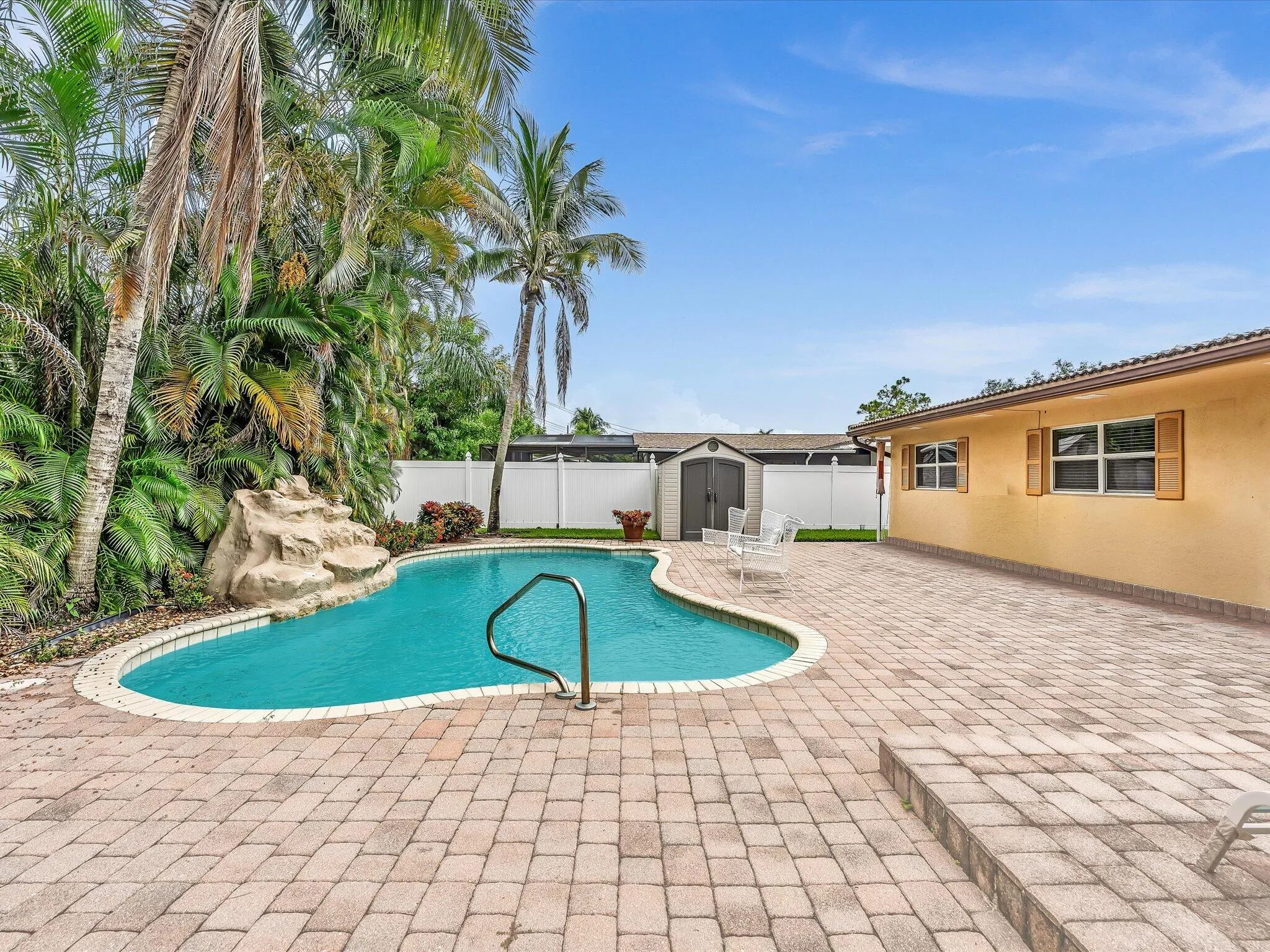Paved patio with a freeform swimming pool, integrated rock feature, and mature tropical landscaping