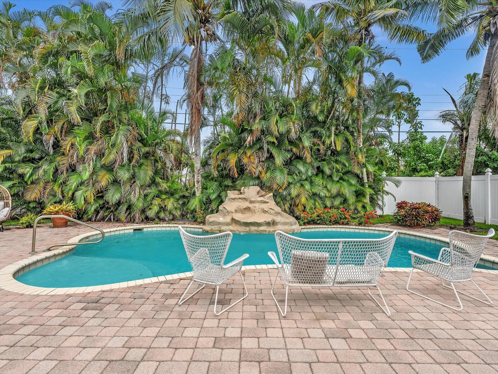 Paved patio area, curved swimming pool with a rock water feature, mature tropical landscaping, and a white privacy fence
