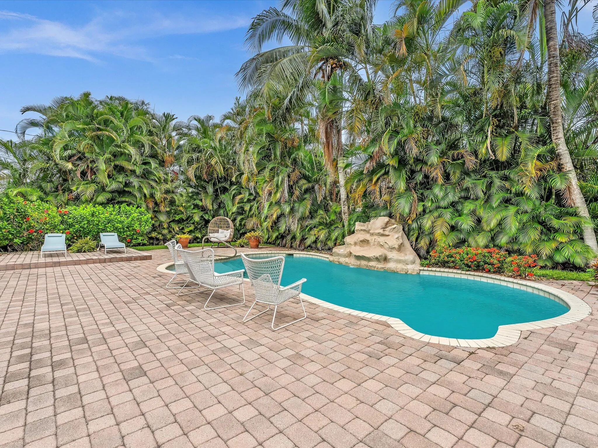 Paved patio featuring a kidney-shaped pool with a rock water feature, surrounded by lush tropical landscaping and mature palm trees