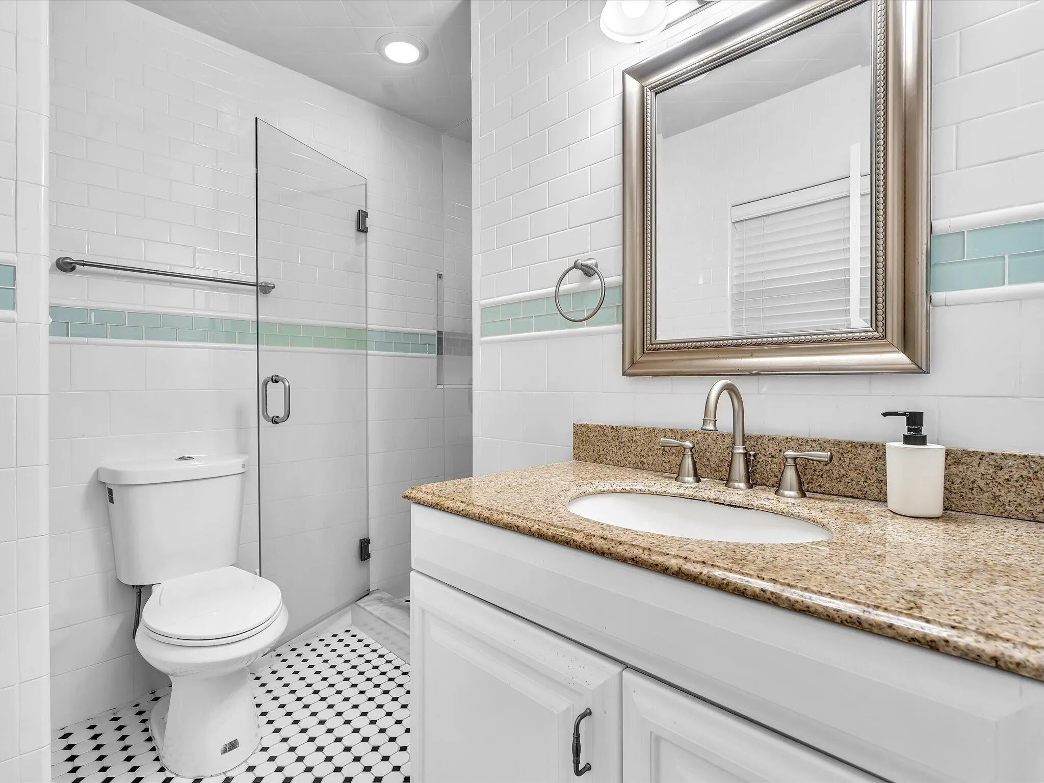 White subway tile walls with an accent tile band, frameless glass shower enclosure, white vanity with an integrated sink, granite countertop, and black and white patterned floor tile