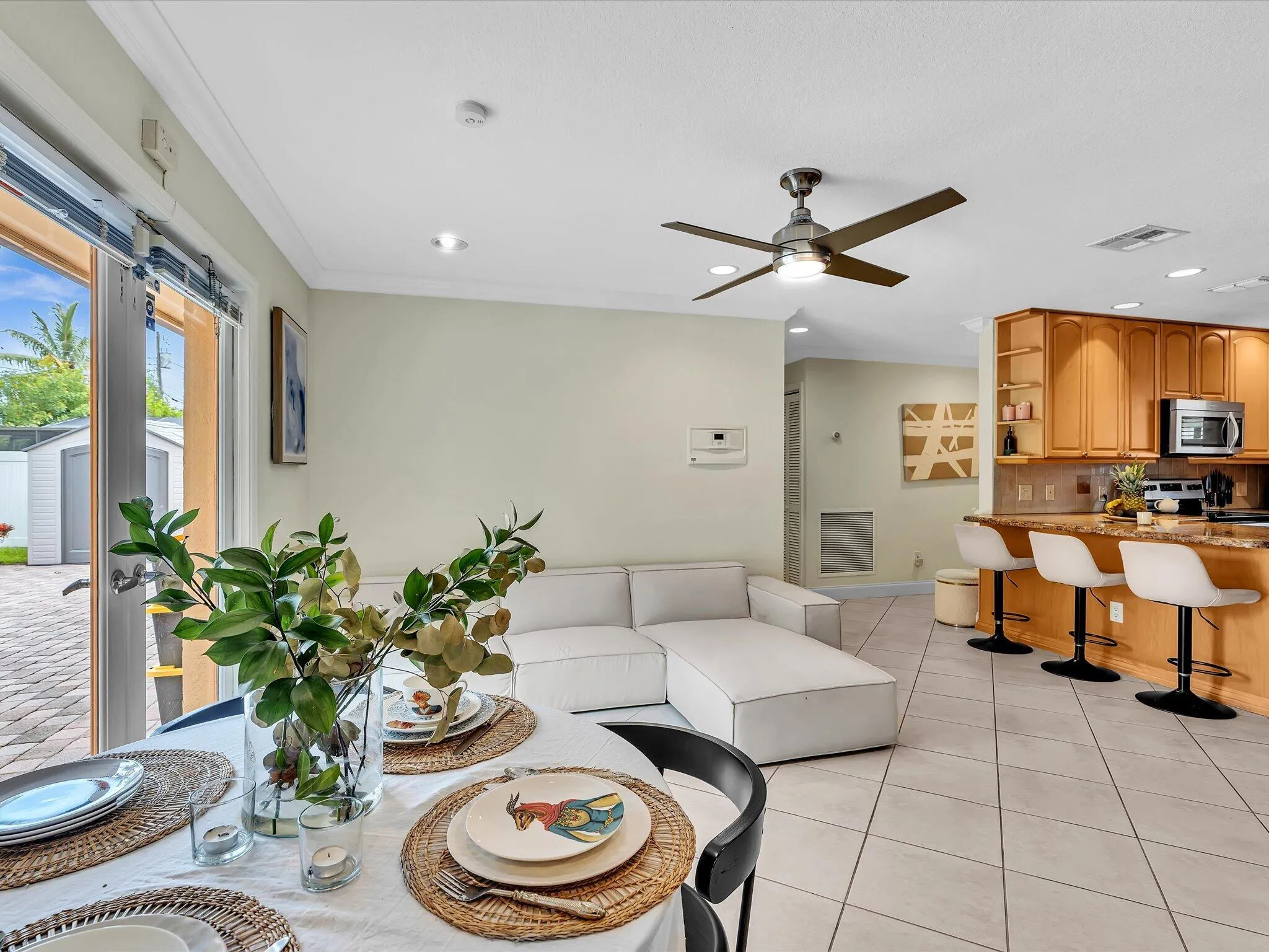 Open-concept dining area with tile flooring and a ceiling fan, featuring a kitchen peninsula with wood cabinetry and built-in appliances, and direct access to an outdoor patio with brick pavers