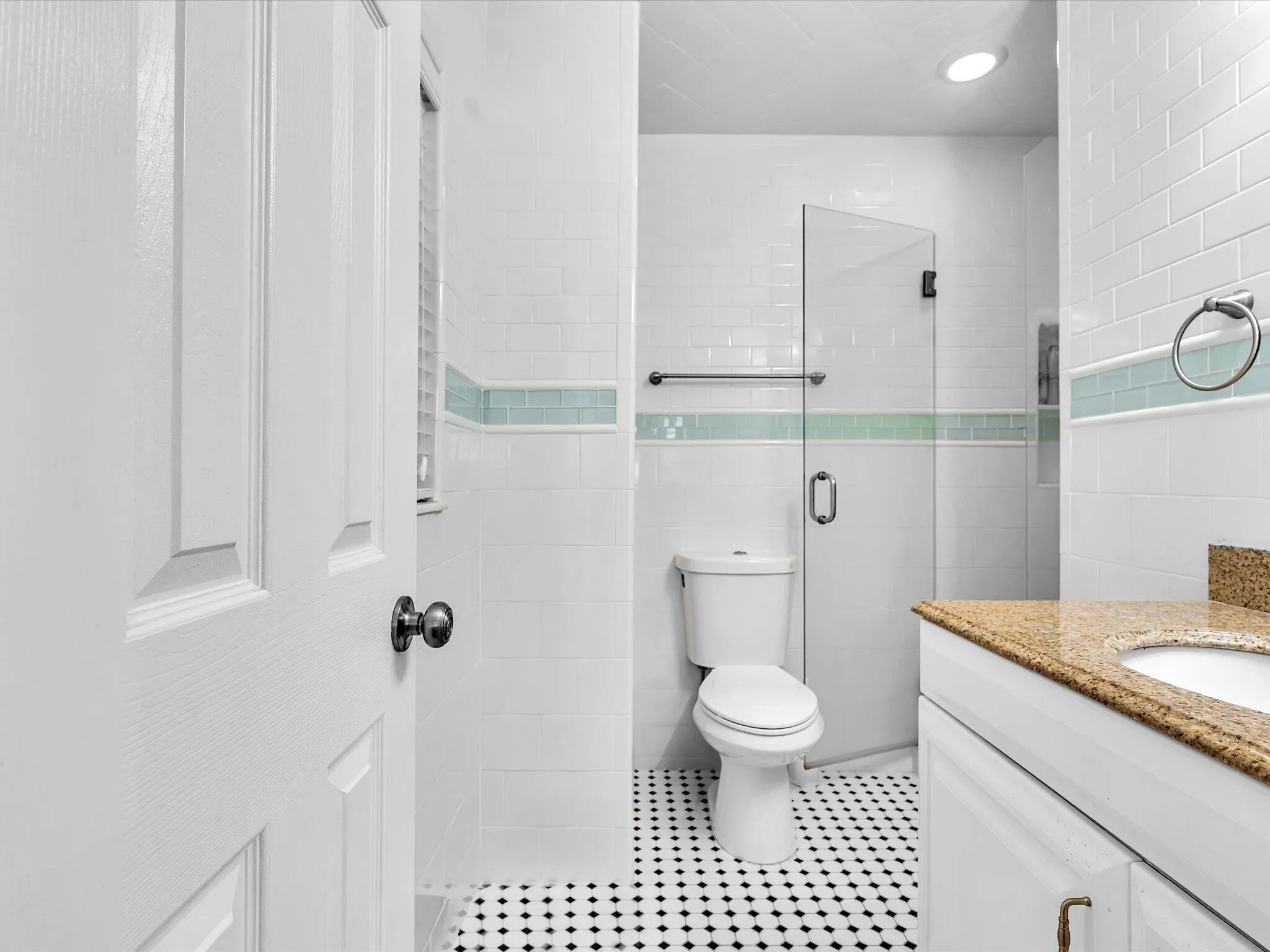 White subway tile walls with a decorative accent band, black and white mosaic floor tile, glass shower enclosure, single vanity with a stone countertop, and a built-in toilet