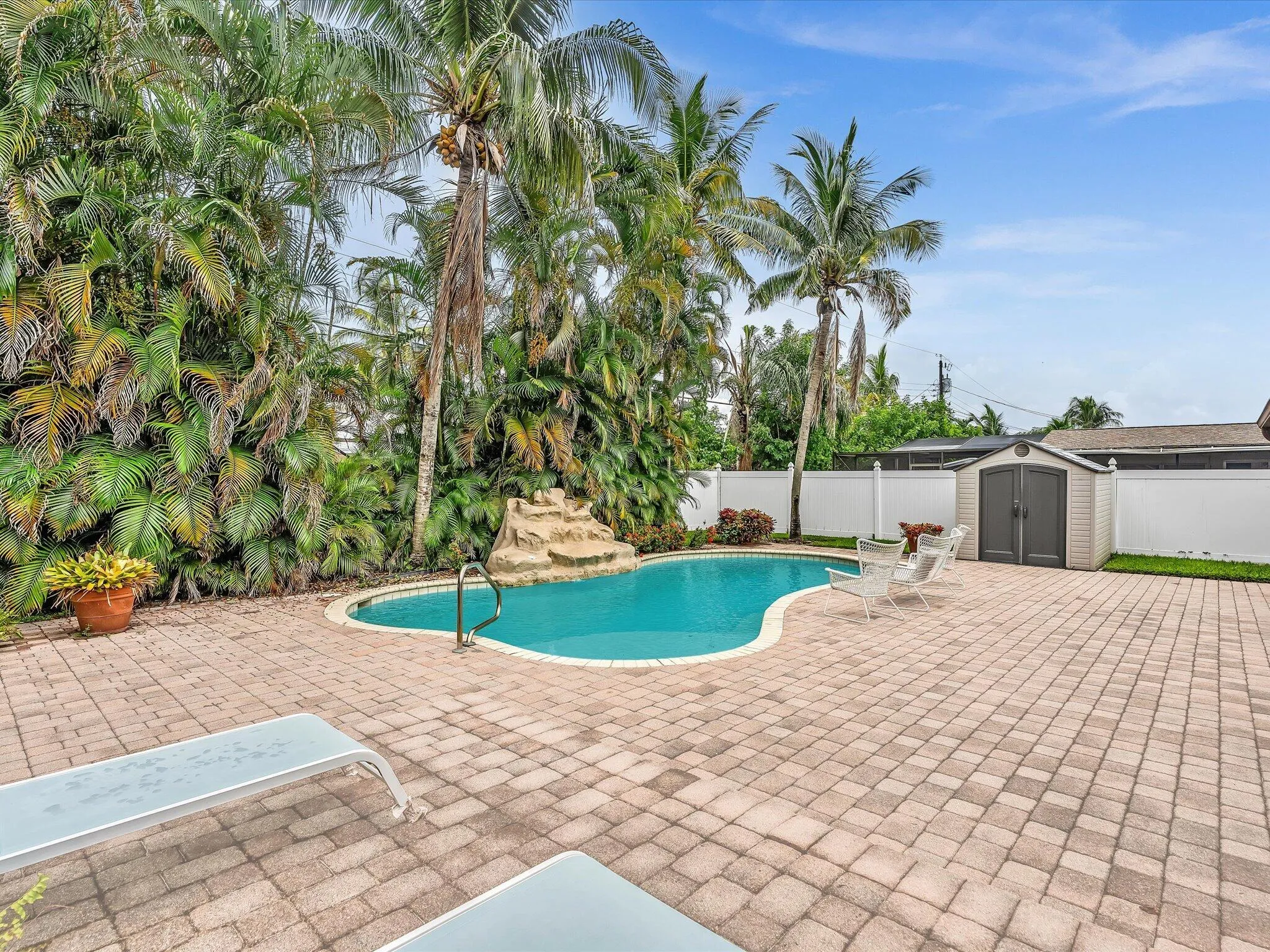 Freeform swimming pool with rock waterfall feature, paver patio, lush tropical landscaping, privacy fencing, and a detached storage shed