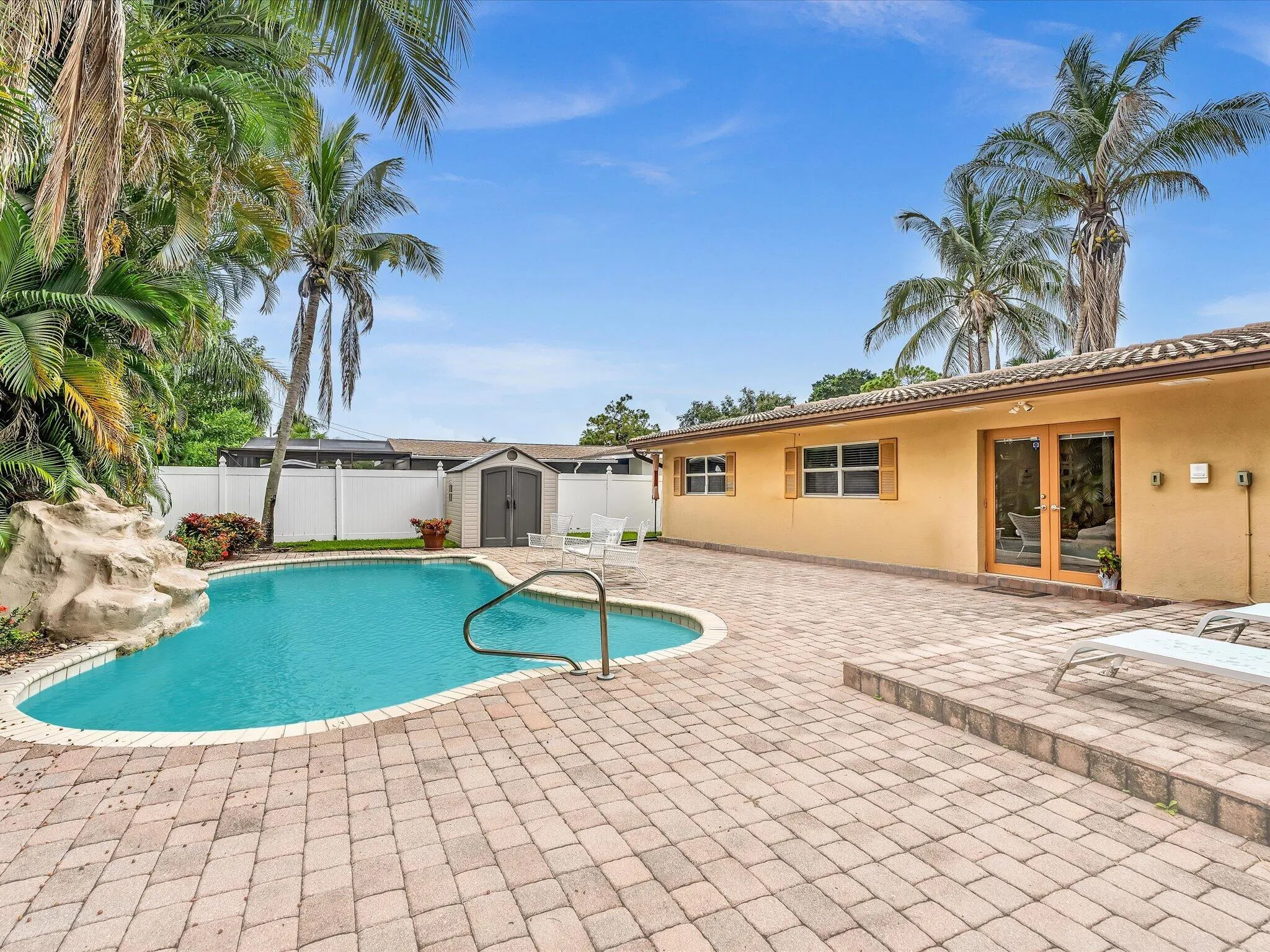 Resort-style backyard oasis featuring a freeform saltwater pool with integrated spa, expansive paver patio, mature palm landscaping, and a single-story residence with yellow stucco exterior and sliding glass doors opening to the outdoor entertaining space
