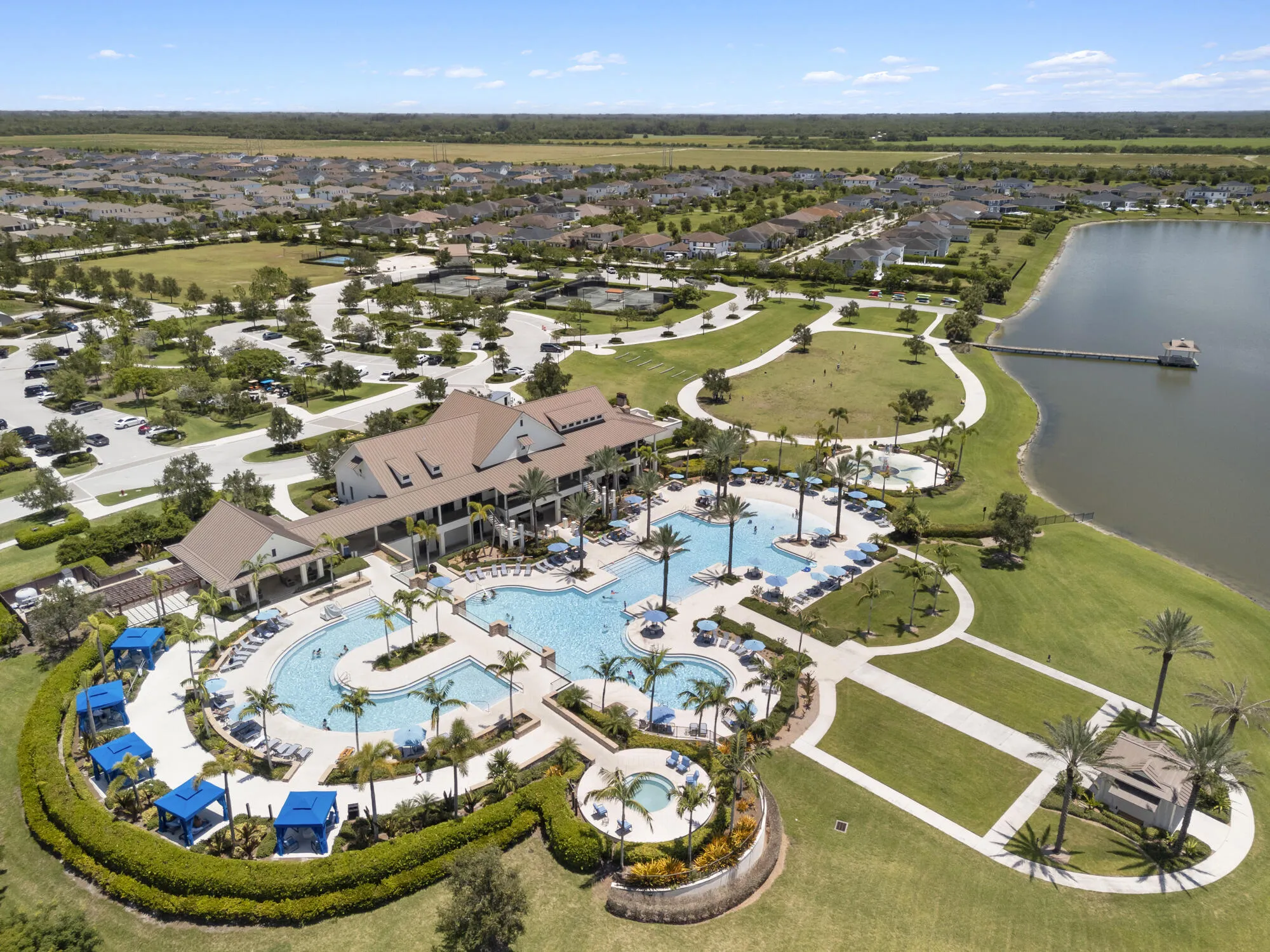 Expansive community pool complex featuring multiple swimming areas, a large clubhouse building, surrounding green spaces, a pier extending onto a body of water, and patterned pathways