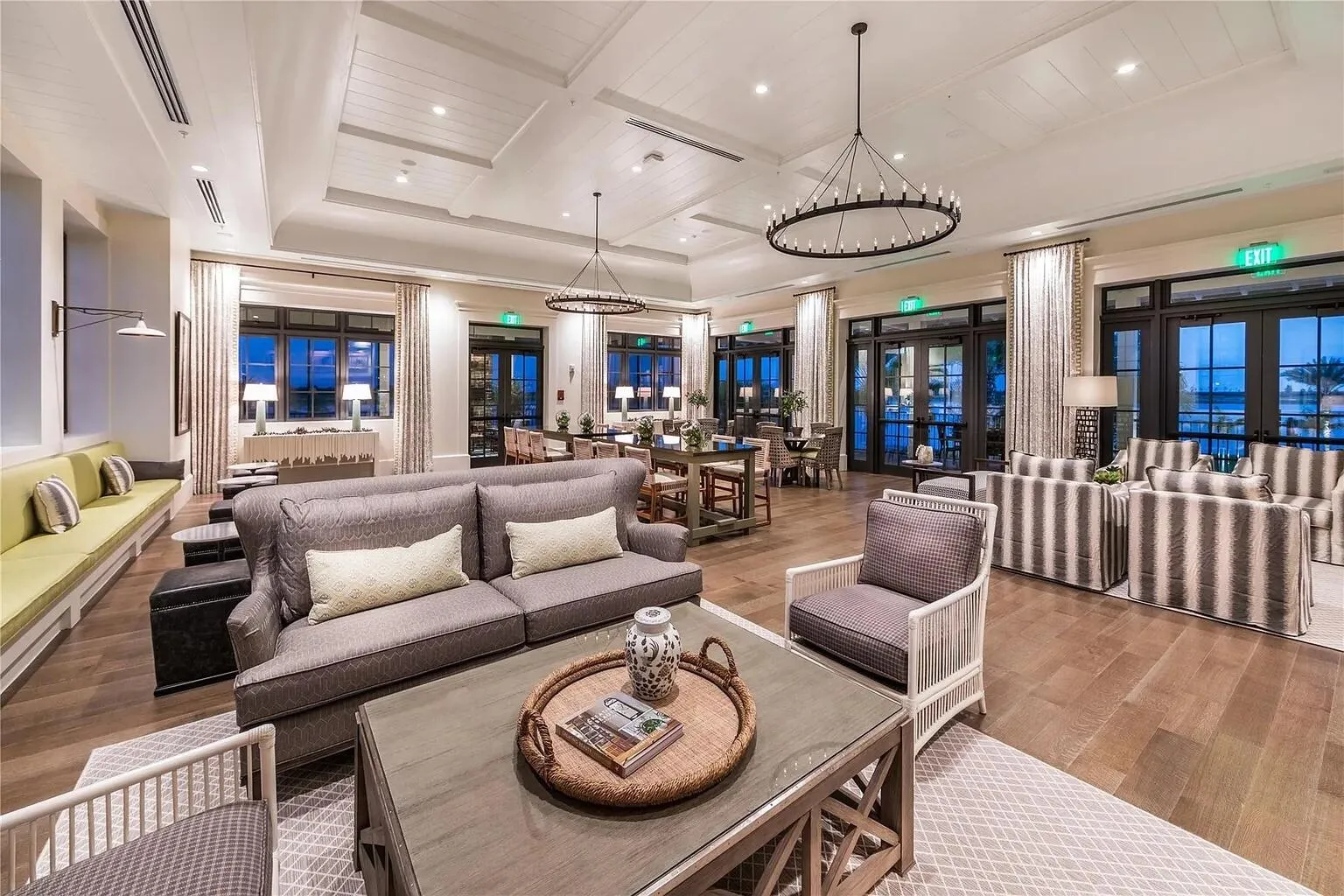 Spacious clubhouse great room with high ceilings, statement chandeliers, plush seating areas, and floor-to-ceiling windows overlooking the community lake at dusk.