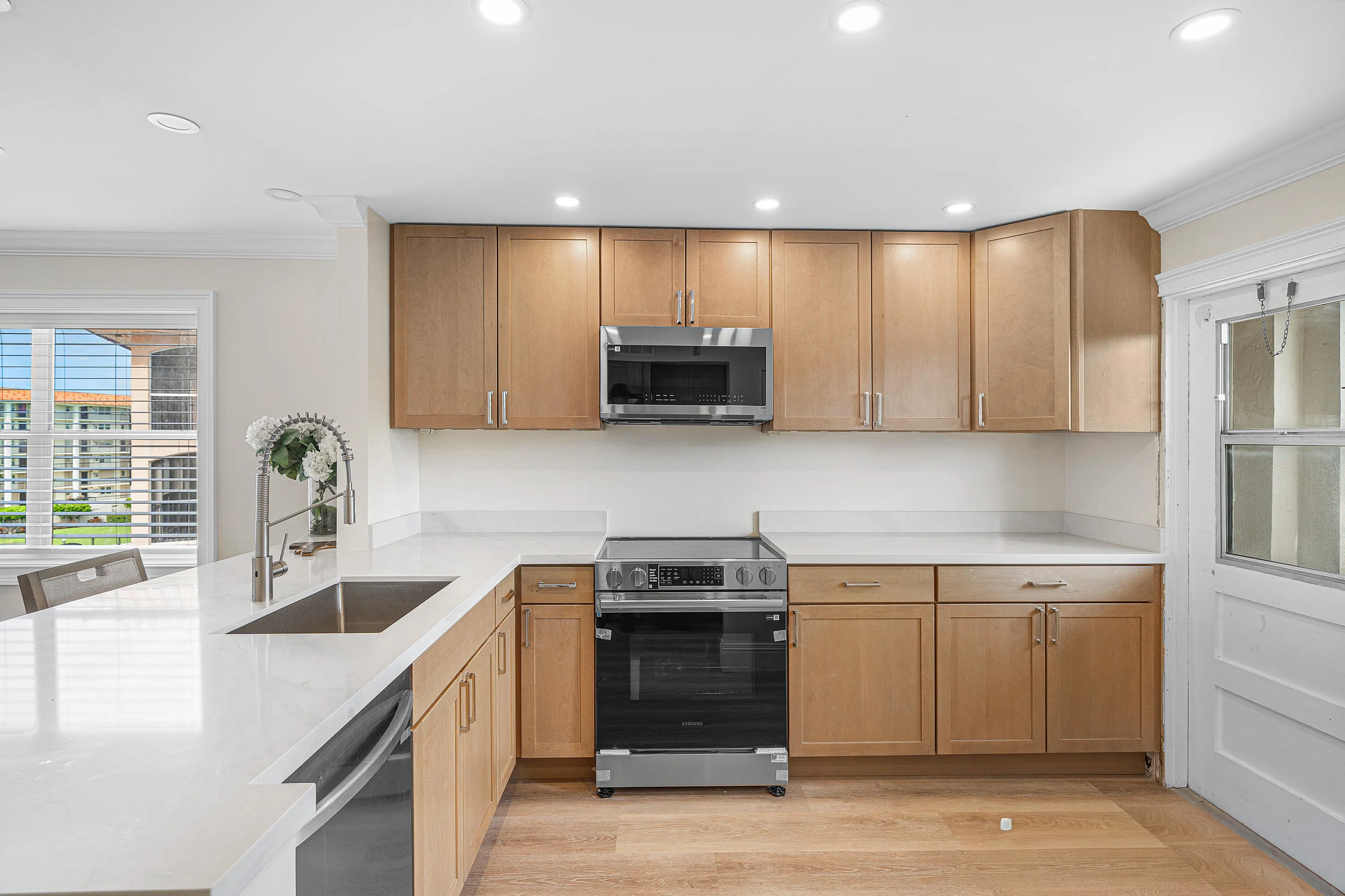 Modern kitchen featuring warm wood-finish cabinetry, stainless steel gas range and microwave, undermount sink with chrome fixtures, white quartz countertops, recessed lighting, and wood-finish flooring throughout the open layout