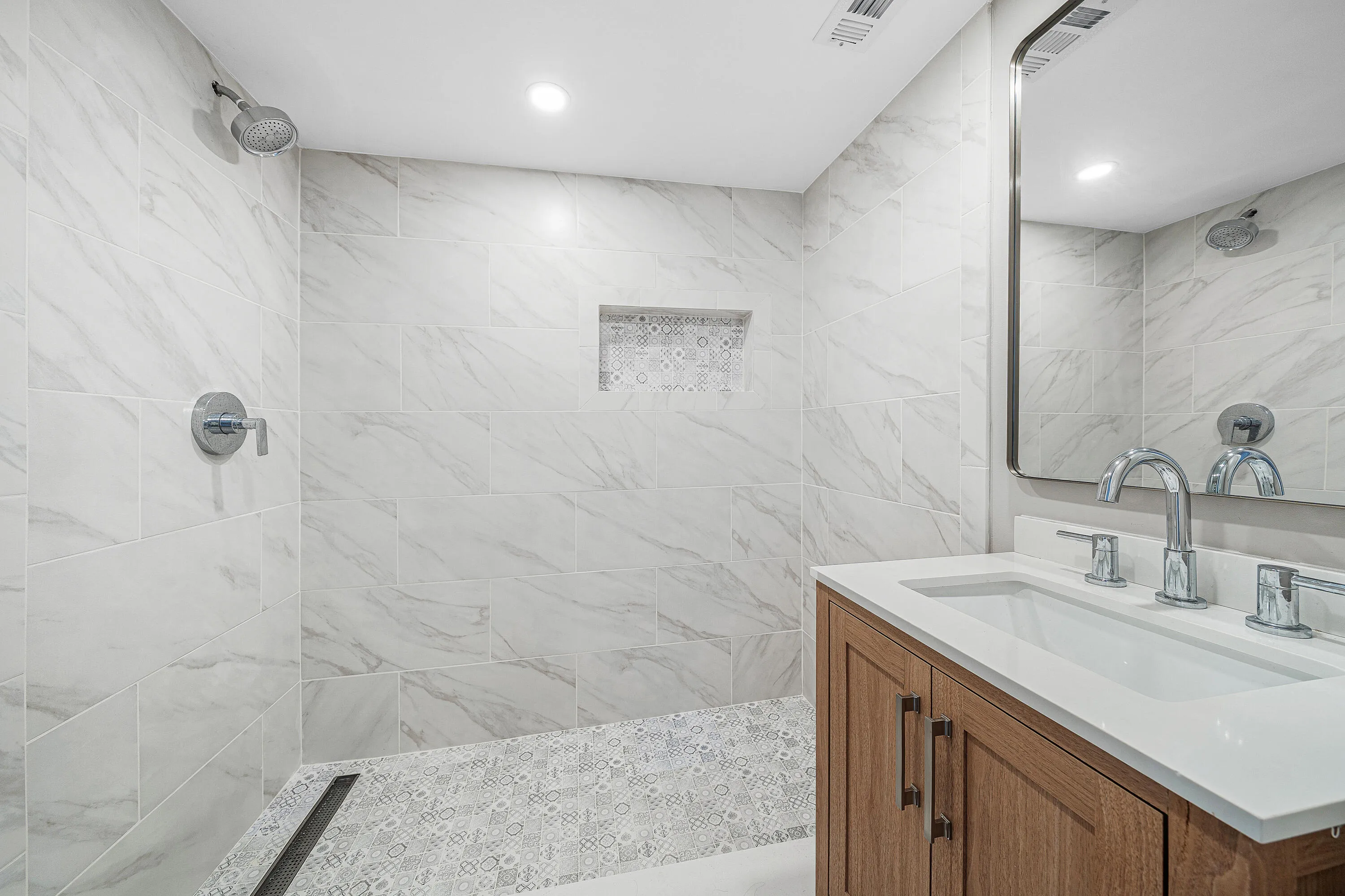 Spa-inspired primary bathroom featuring marble-look tile walls and flooring, a walk-in shower with frameless glass enclosure and rainfall showerhead, recessed ceiling lighting, and a wood-finish vanity with white quartz countertop and chrome fixtures