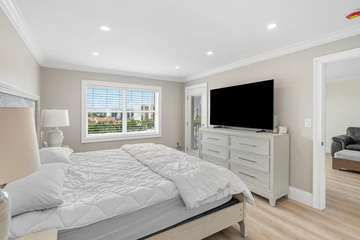 Spacious primary bedroom featuring recessed ceiling lighting, a large window with plantation shutters overlooking landscaped grounds, light wood-finish flooring, and a white painted dresser with ample storage