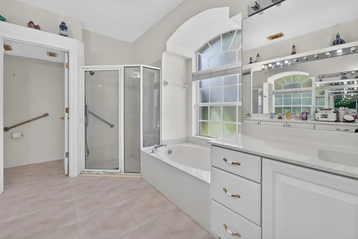 Tiled flooring, corner soaking tub, glass-enclosed shower, dual vanity with integrated sinks, and an arched window with privacy blinds