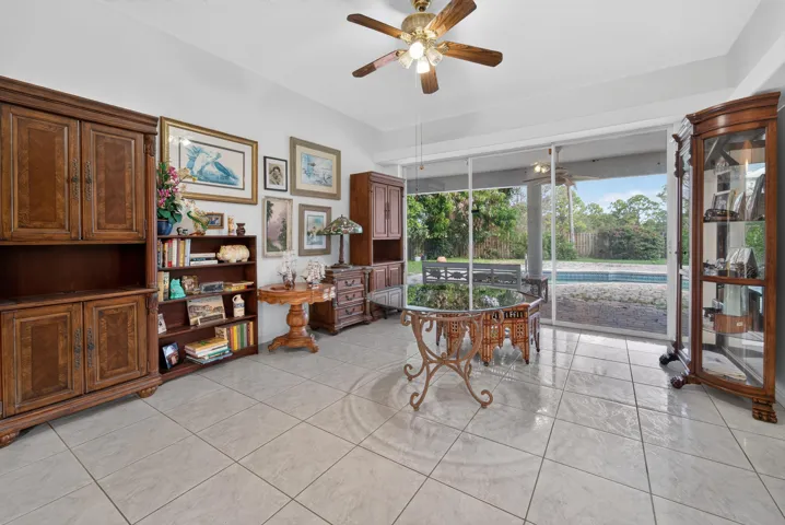 Bright, light-filled den featuring tile flooring, wood cabinetry with glass-front display cases, and a ceiling fan with integrated lighting