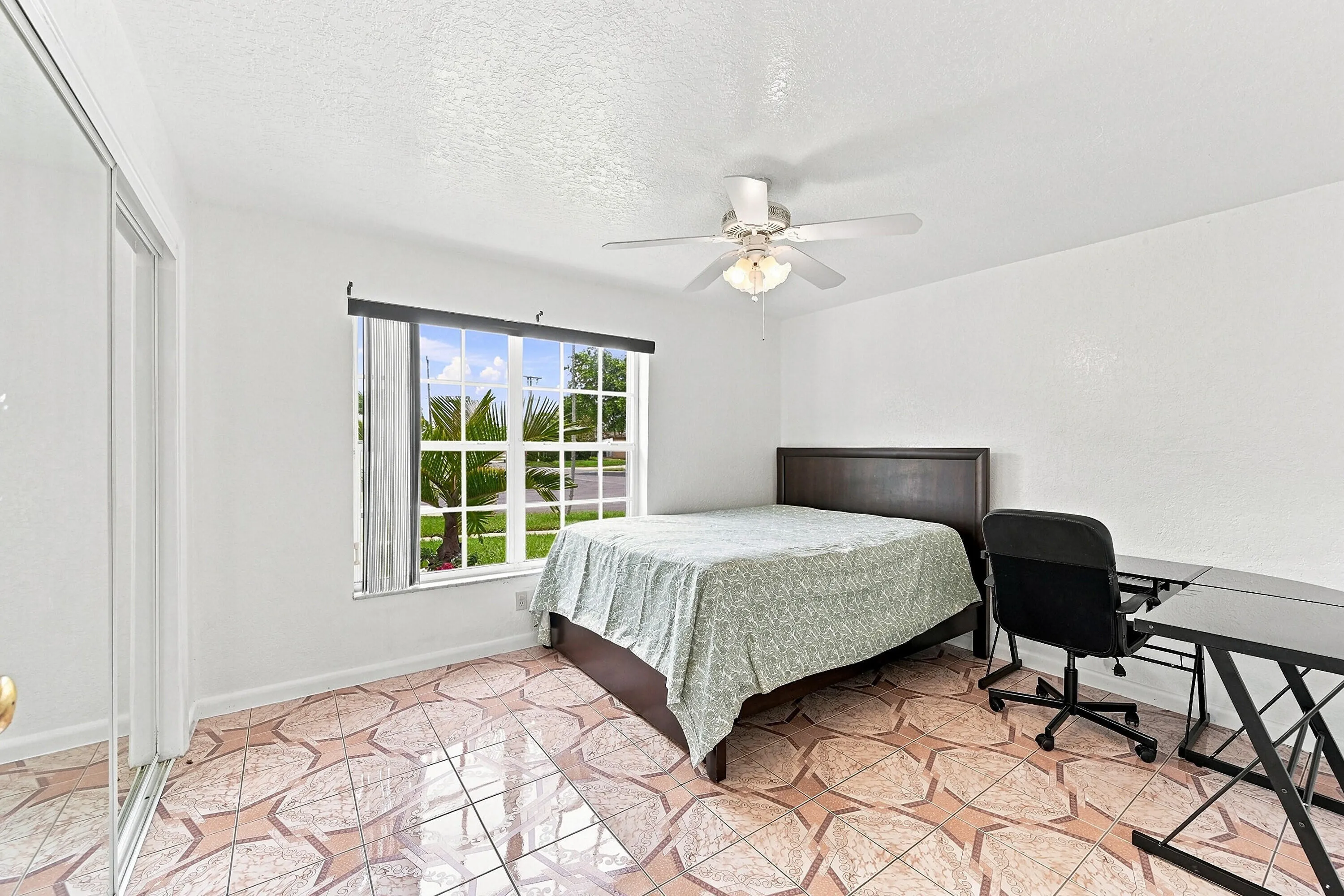 Spacious bedroom featuring geometric patterned tile flooring, a ceiling fan with integrated lighting, large multi-pane windows with vertical blinds and sheer curtains providing natural light, and a dark wood-finish bed frame
