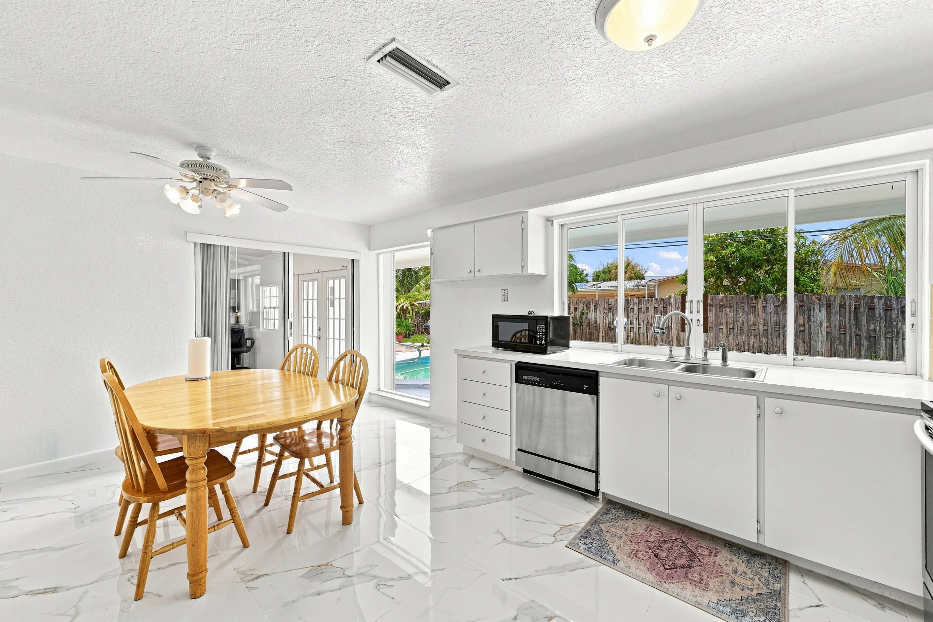 Bright, open-concept kitchen and dining area featuring white cabinetry with stainless steel appliances, marble-finish flooring throughout, a wooden dining table with seating for four, ceiling fan with integrated lighting, and expansive windows overlooking the backyard with mature landscaping and pool access