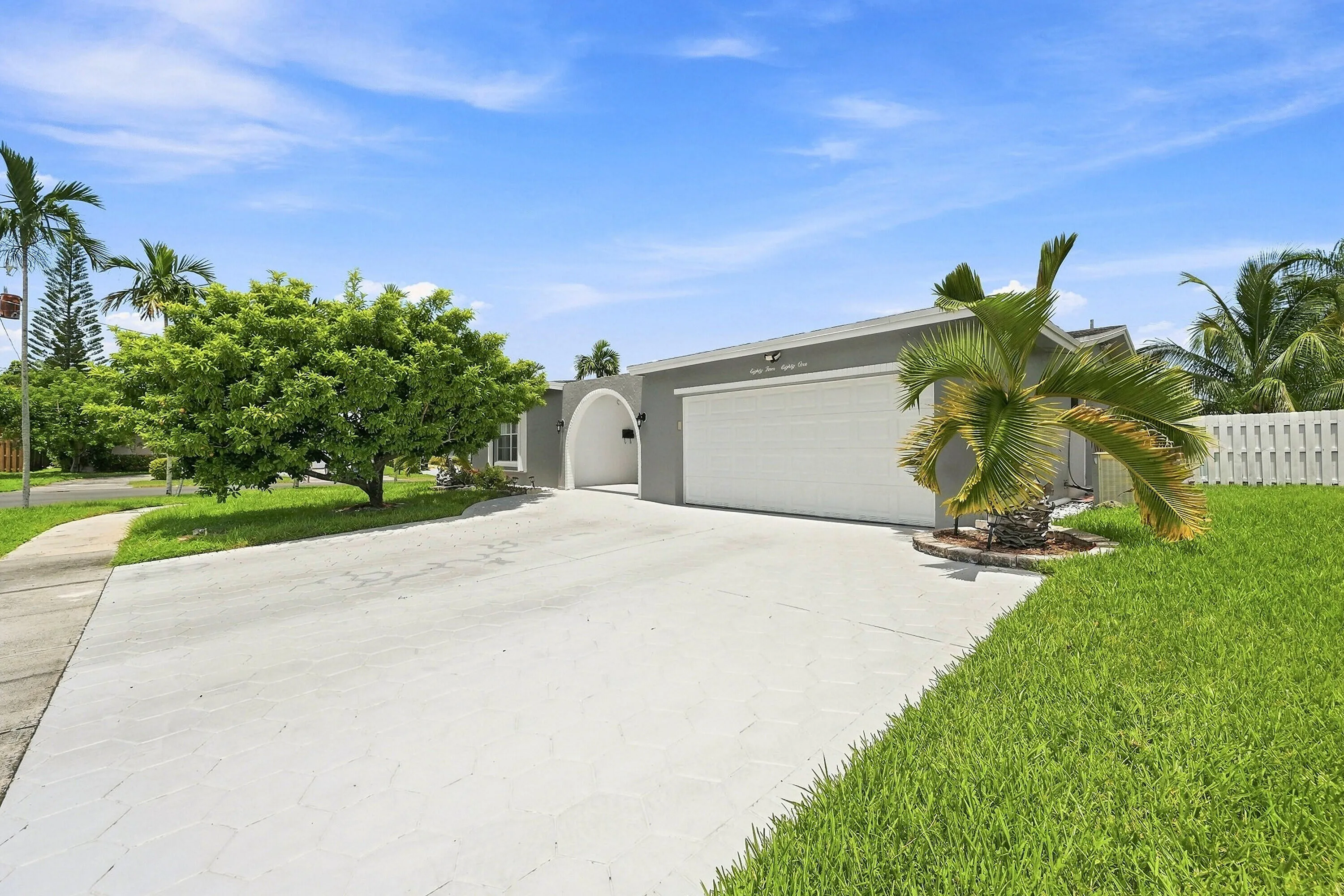 Welcoming front elevation featuring a spacious concrete driveway, two-car garage with panel doors, lush manicured lawn, mature landscaping with palm trees and established greenery, and a white picket fence accent along the property perimeter