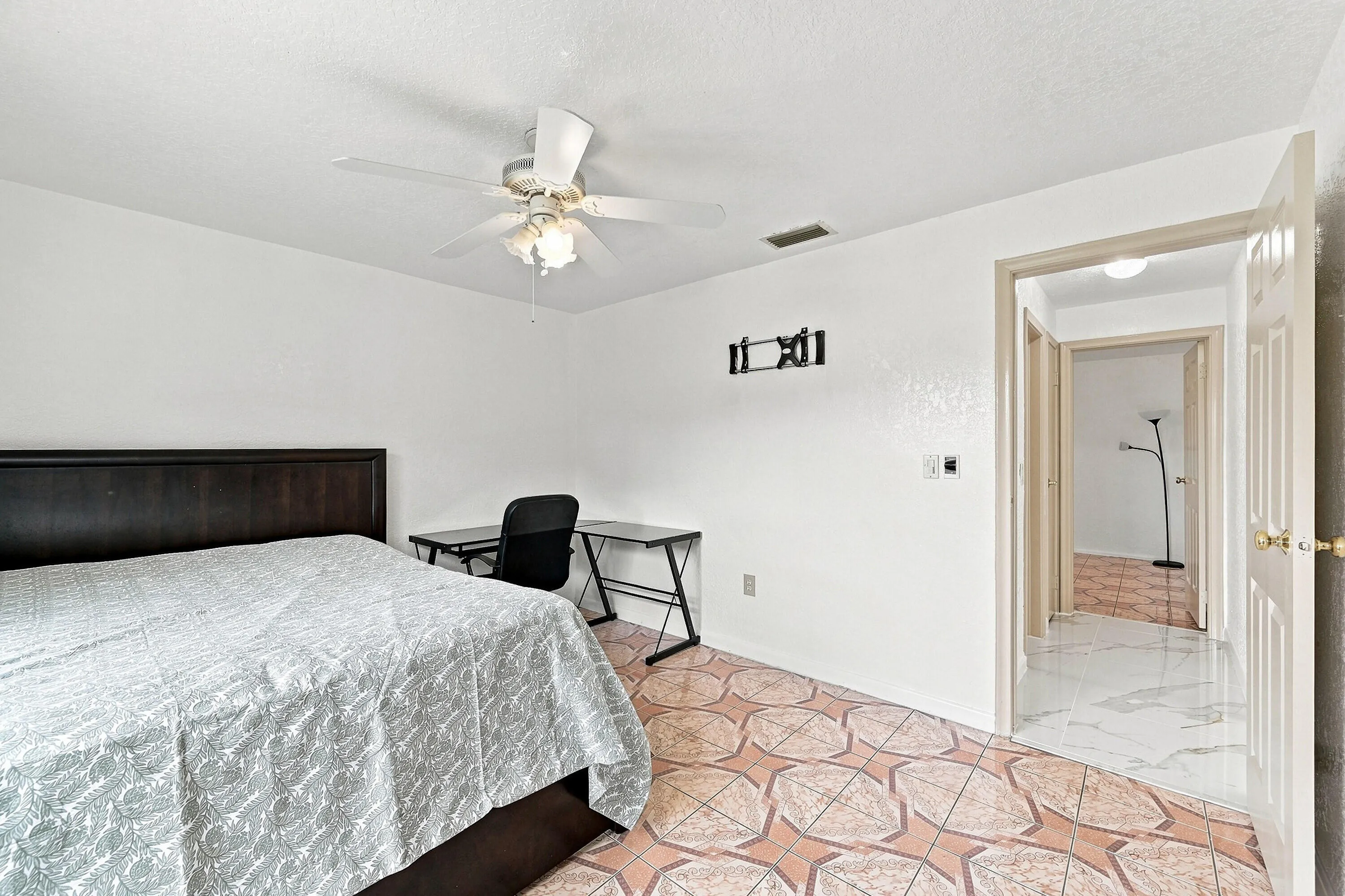 Spacious bedroom featuring a black upholstered bed frame, geometric patterned area rug in warm terracotta tones, ceiling fan with integrated lighting, dedicated workspace with desk and chair, and direct access to an en-suite bathroom through a doorway