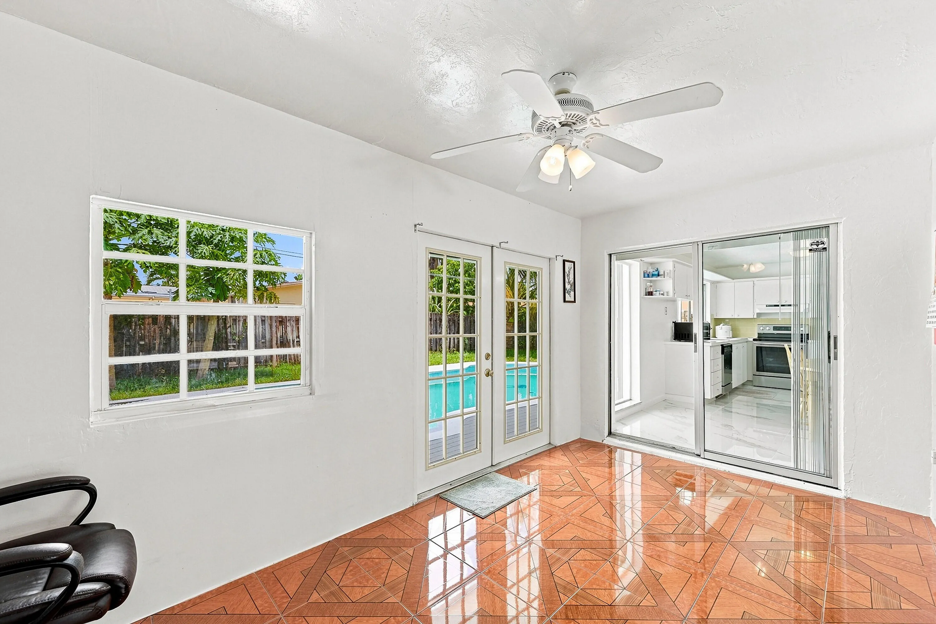 Bright and airy room featuring gleaming wood-finish flooring, abundant natural light from a large multi-pane window, glass sliding doors opening to outdoor space, ceiling fan with integrated lighting, and access to adjacent areas