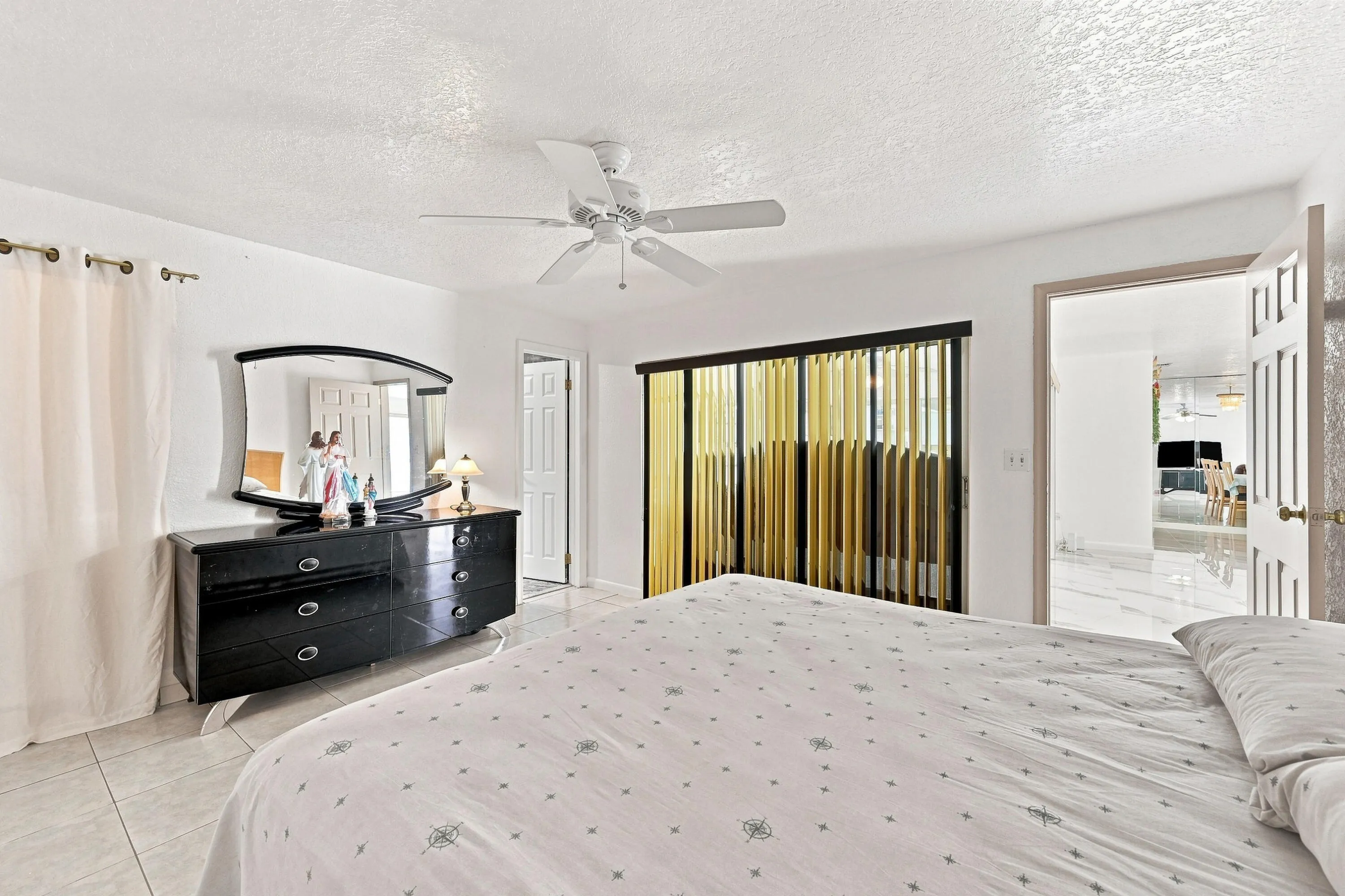 Spacious primary bedroom featuring a ceiling fan, dual nightstands with attached lighting, black wood dresser with mirror, sliding glass doors with yellow vertical blinds, and tile flooring throughout