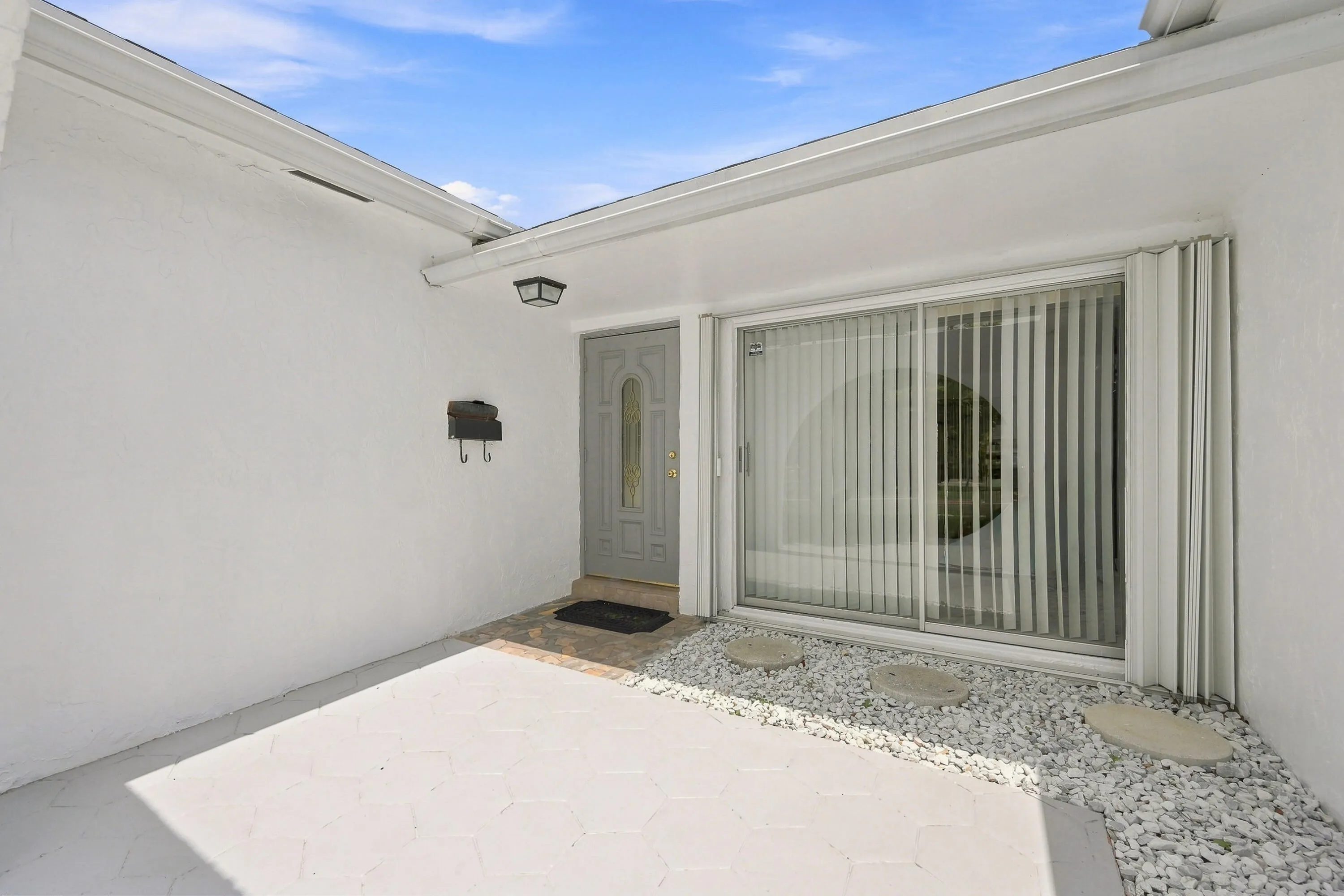 Covered outdoor entertaining space featuring a clean concrete patio, vertical blinds on sliding glass doors, and decorative gravel landscaping