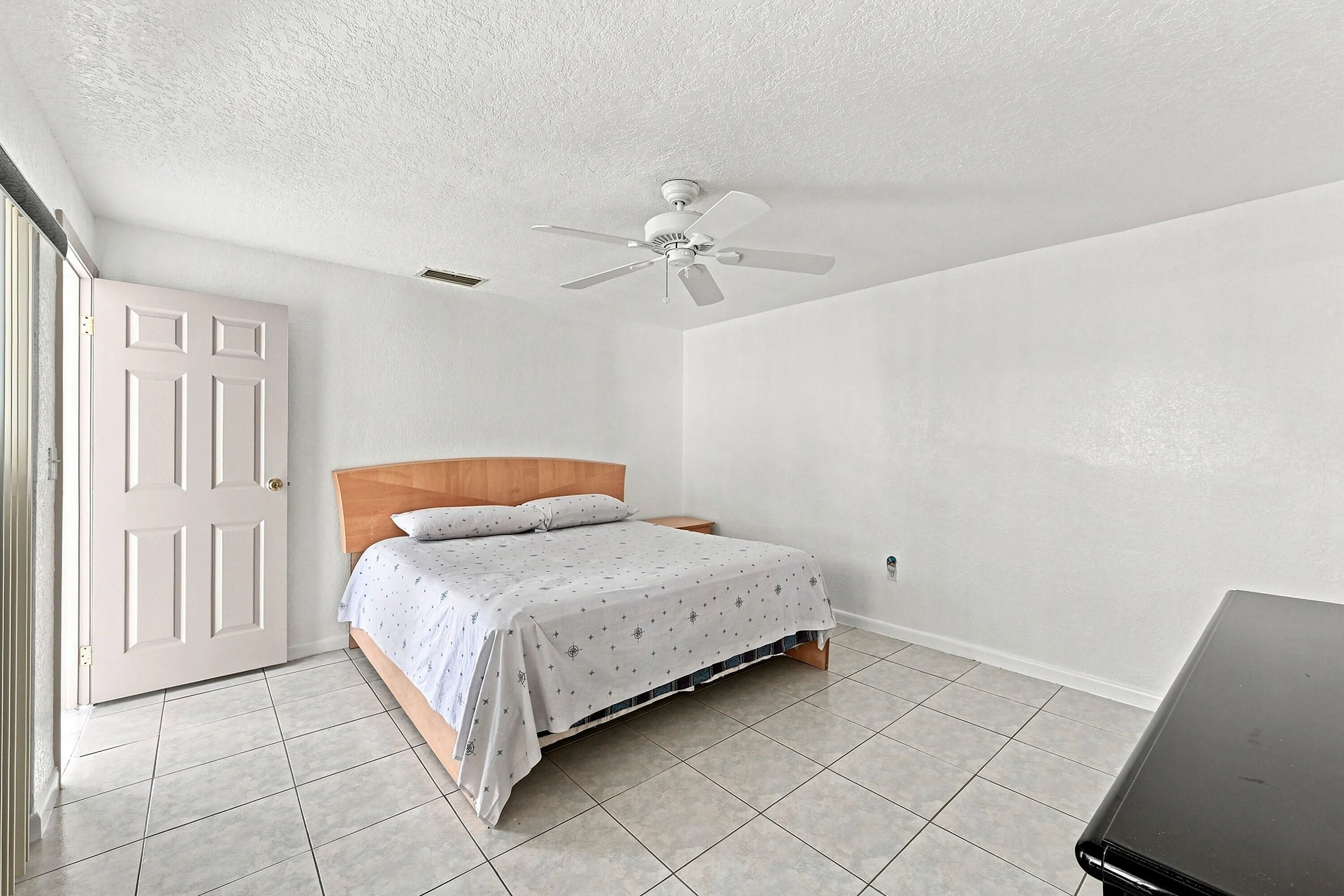 Spacious bedroom featuring a wood-finish platform bed frame, tile flooring throughout, ceiling fan with integrated lighting, and a six-panel closet door