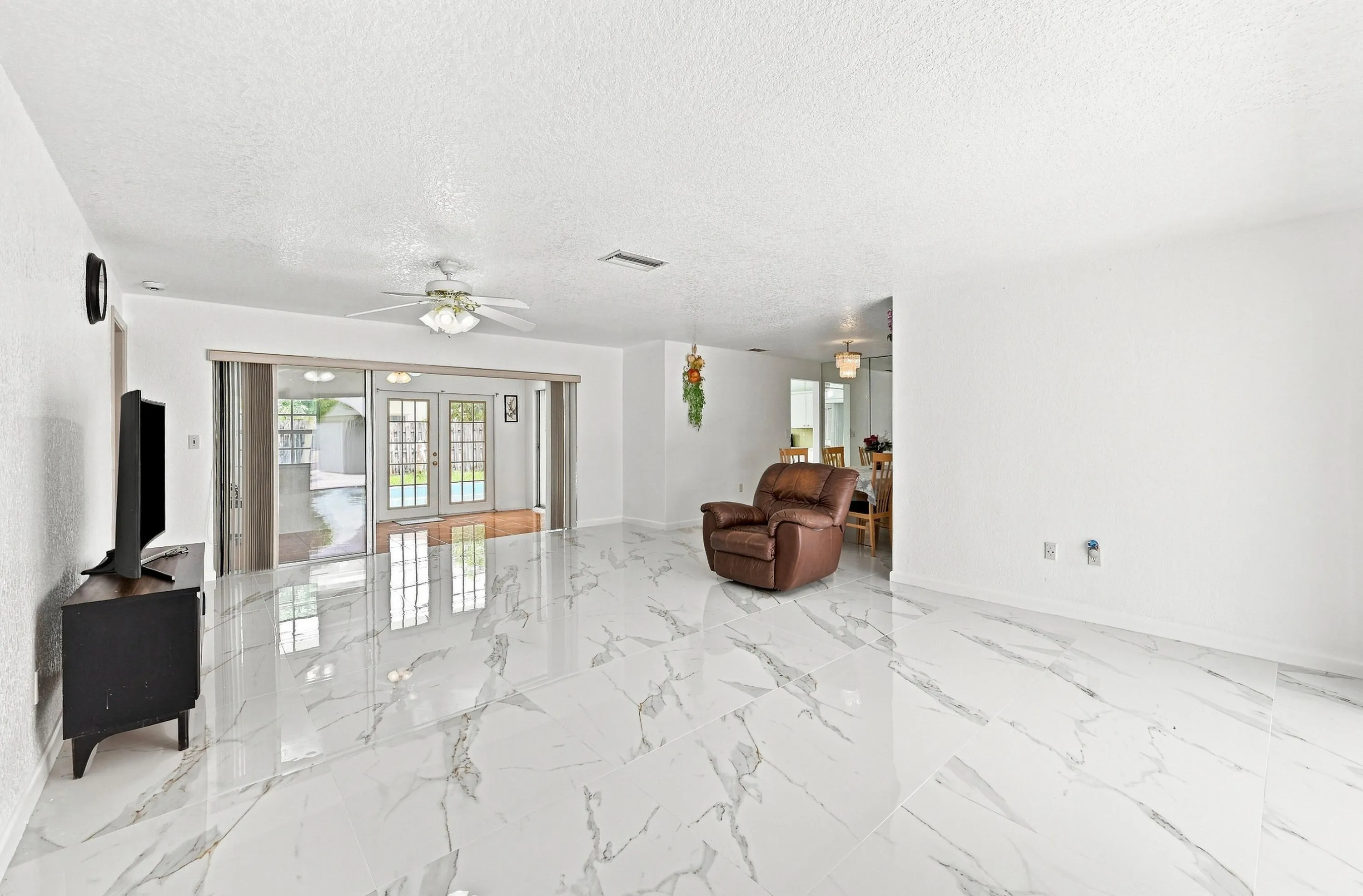 Spacious open-concept living area showcasing elegant veined stone-finish flooring, abundant natural light from French doors and transom windows, ceiling fan with integrated lighting, and seamless sightlines to adjoining rooms