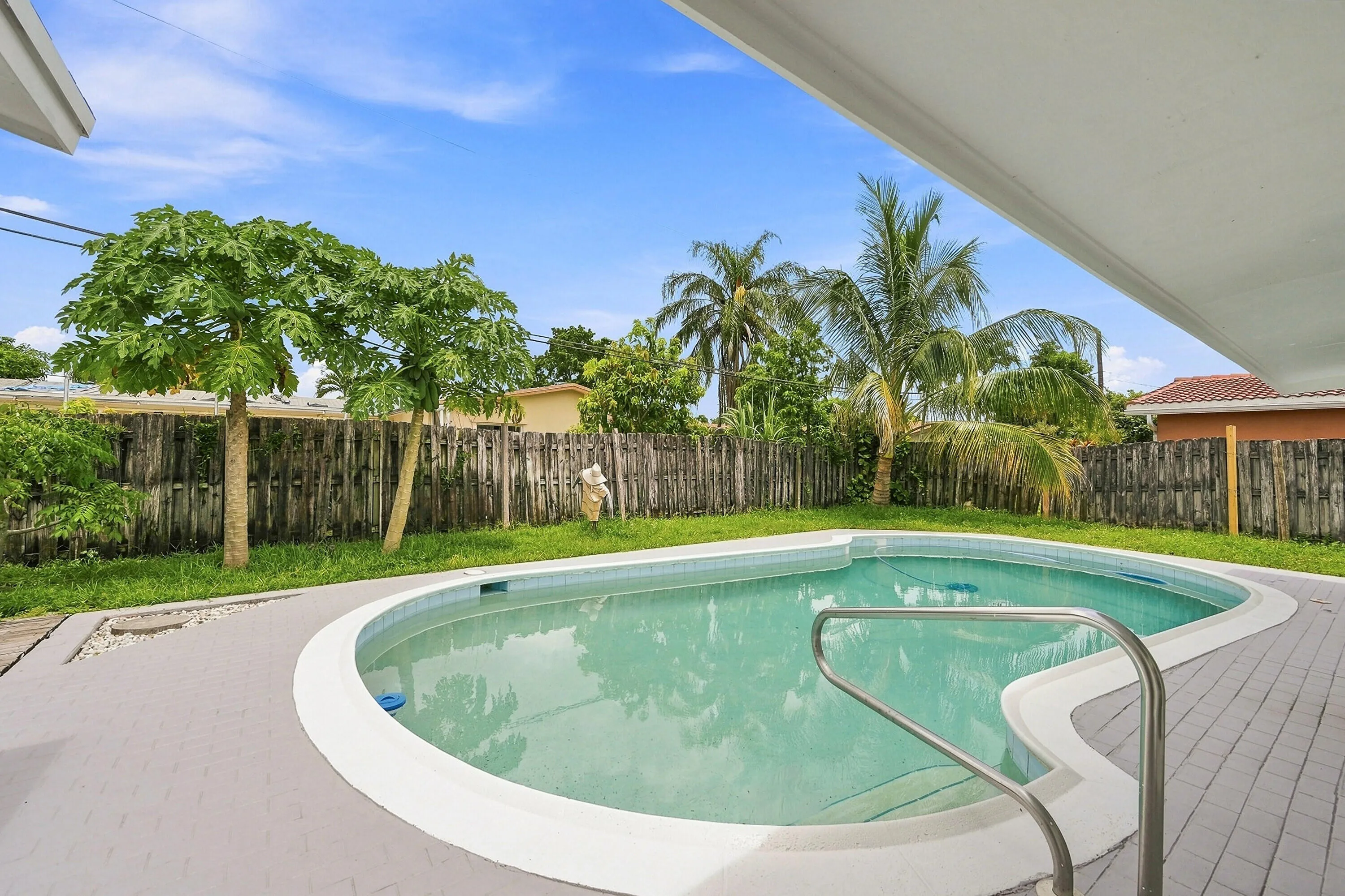 Freeform swimming pool with a handrail, surrounded by a gray paved deck and a wooden privacy fence