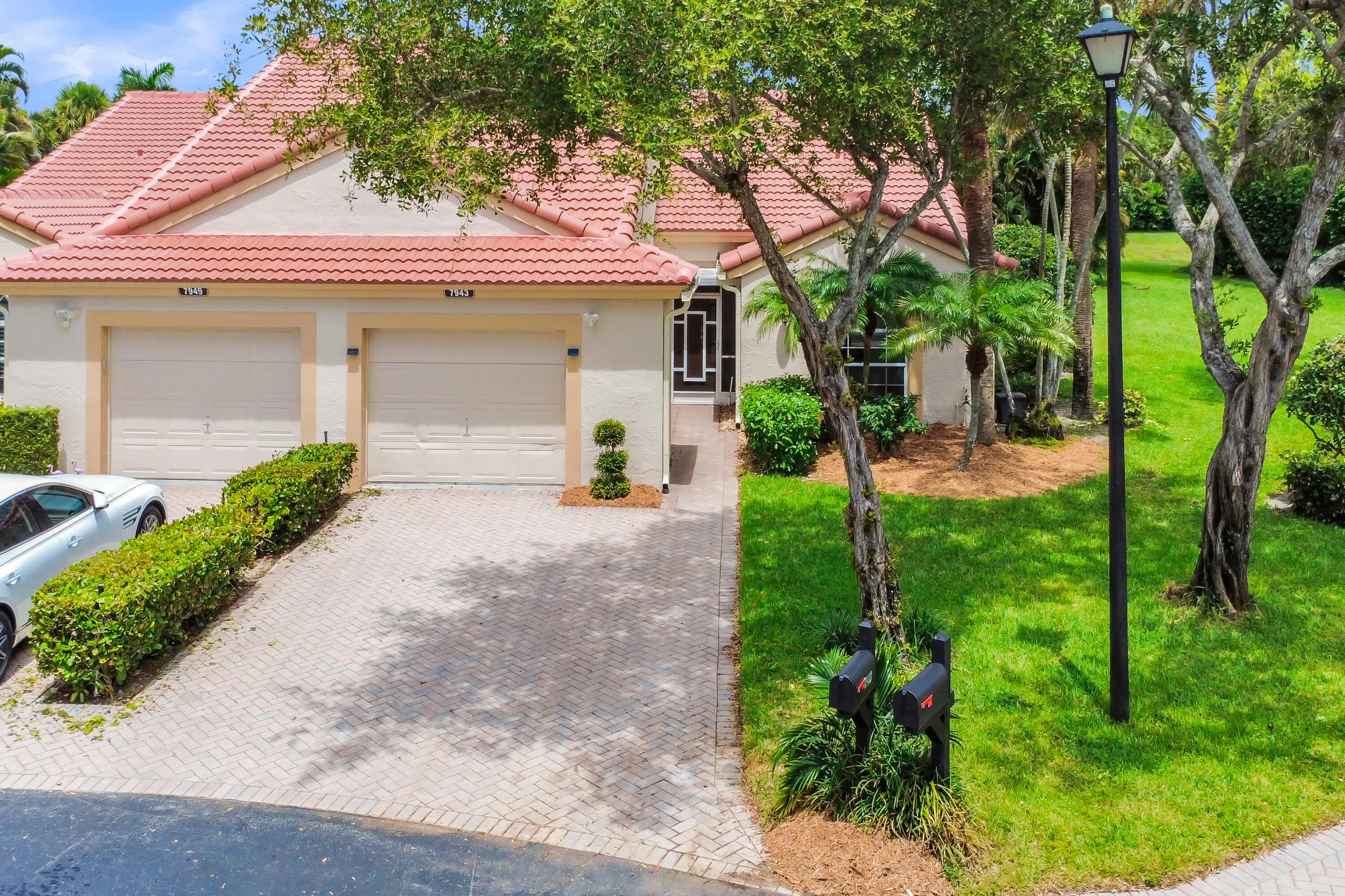 Residential exterior featuring a tile roof, dual garage doors, a paver driveway, and a landscaped entry with mature trees and shrubbery