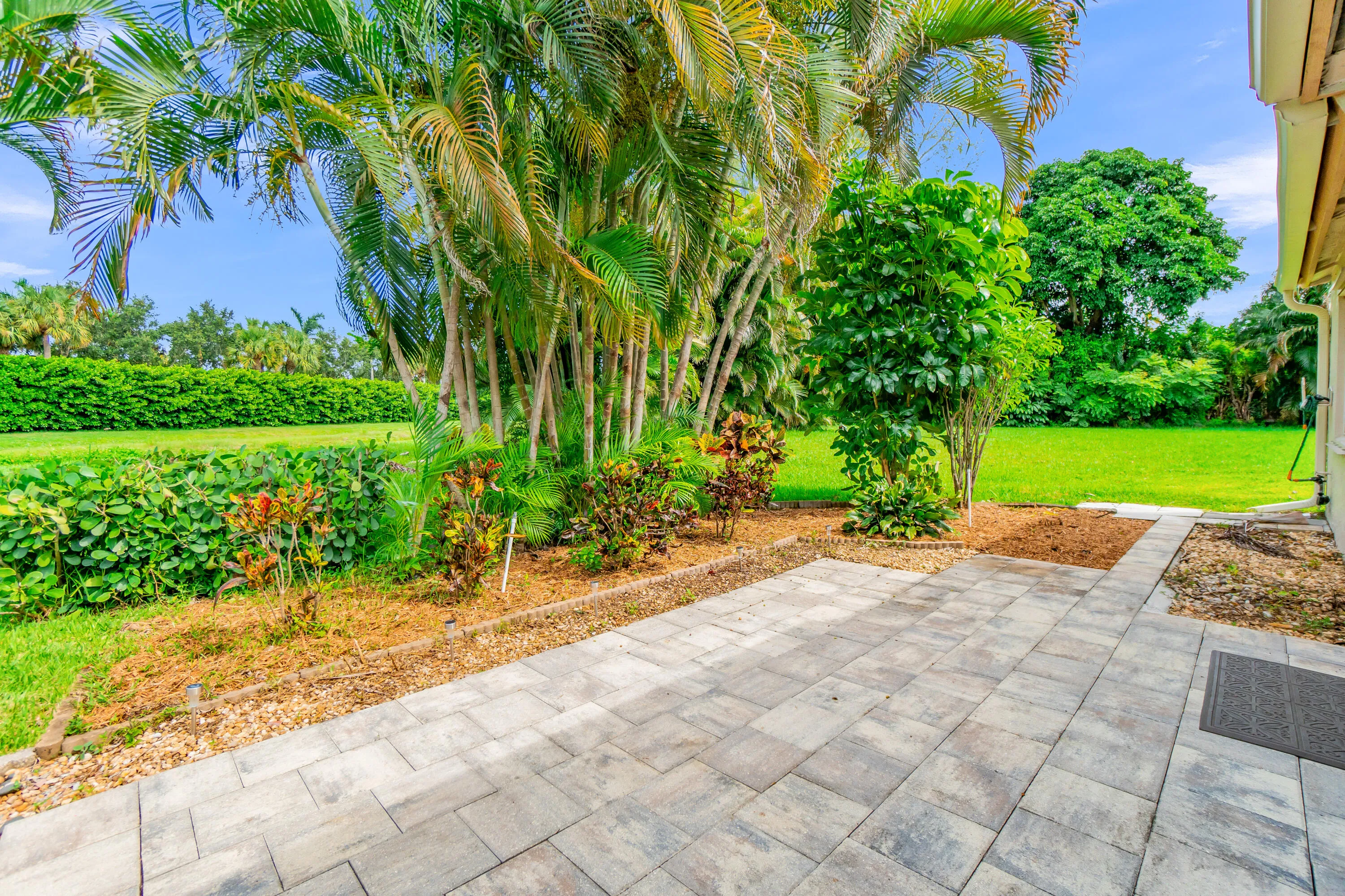 Paver patio area with a landscaped border featuring palm trees, various plantings, and a manicured lawn