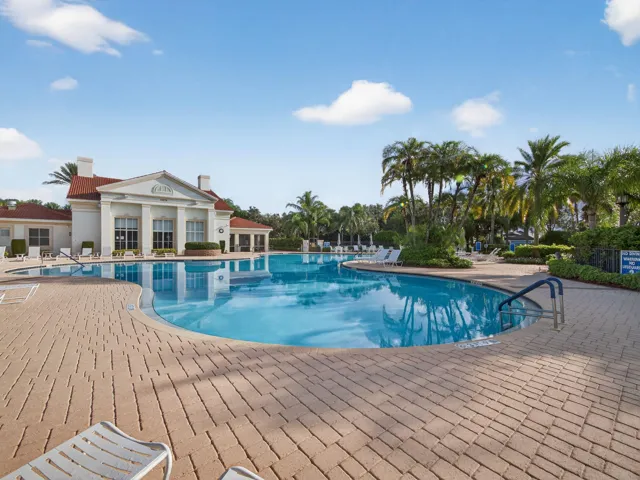 Large outdoor swimming pool with surrounding brick paver deck, lush tropical landscaping, and a classical-style clubhouse building