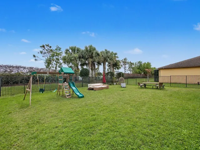 Expansive grounds featuring a lush, level lawn with mature palm and shade trees, children's play equipment with slide and swing set, sandbox area, and a metal-frame outbuilding