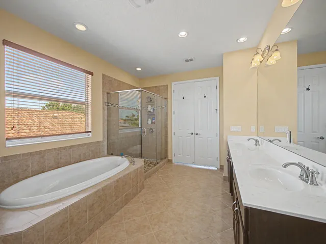 Spa-inspired master bathroom featuring a soaking tub with tile surround, frameless glass-enclosed shower, dual vanity with white cabinetry and marble-finish countertops, recessed lighting, and abundant natural light from large window with horizontal blinds