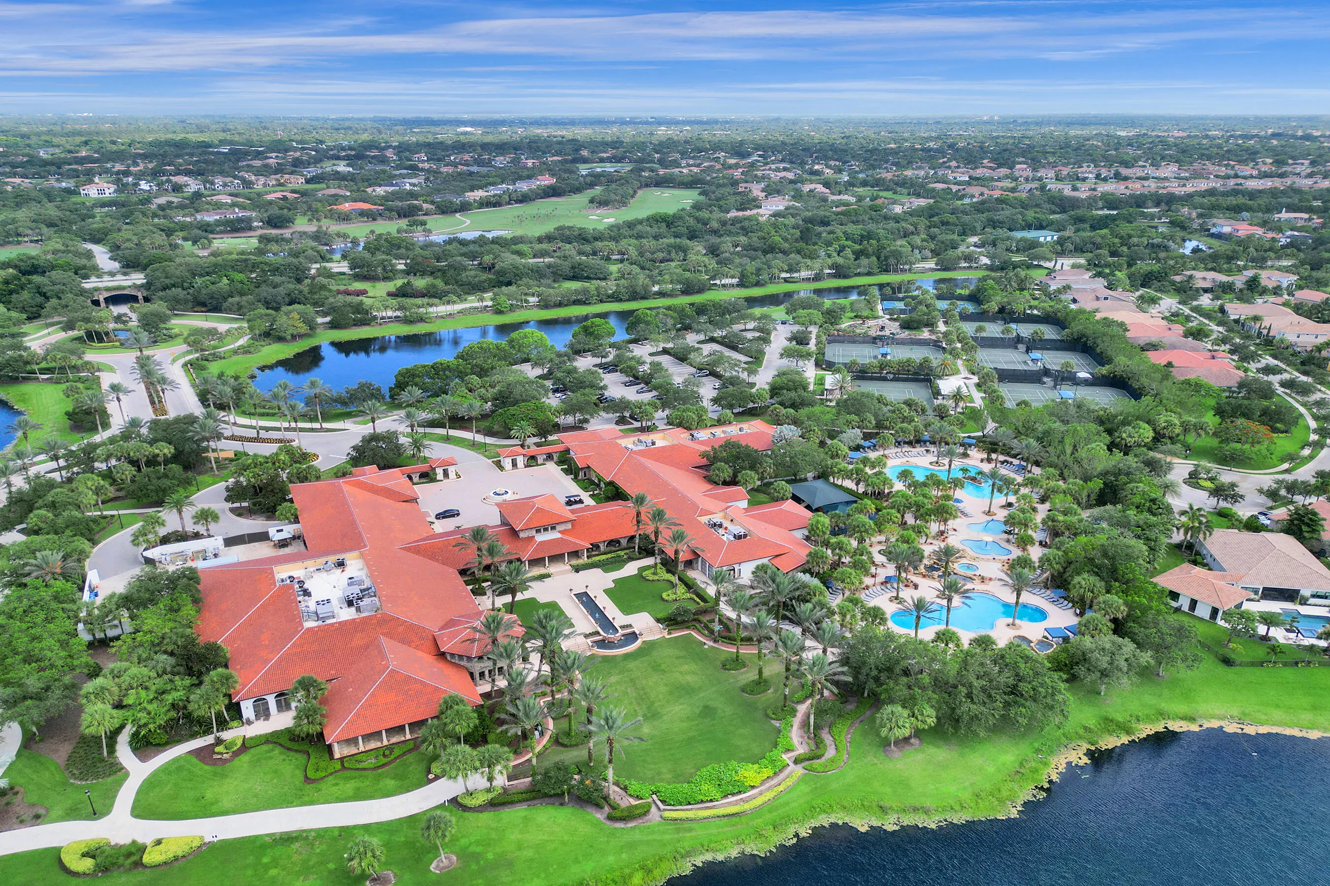 Sprawling property featuring multiple buildings with red tile roofs, multiple swimming pools, several tennis courts, and expansive green spaces with mature trees and water features